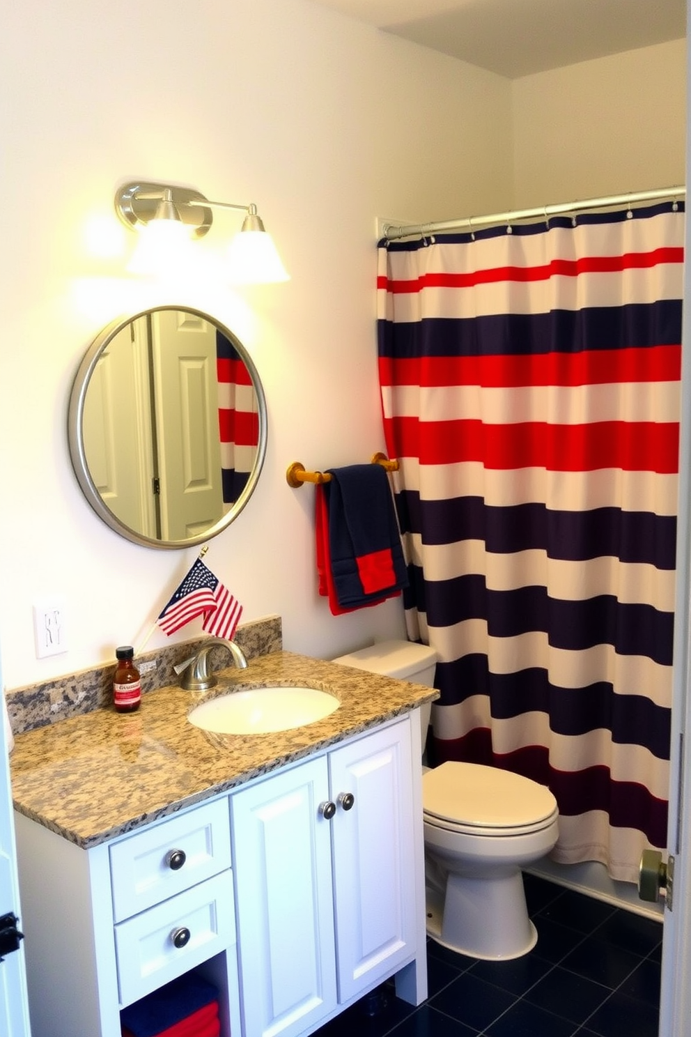 A cozy bathroom decorated for Memorial Day with a red and blue striped shower curtain as the focal point. The walls are painted in a soft white, and the floor is covered with navy blue tiles. A white vanity with a granite countertop supports a single sink, above which hangs a circular mirror with a silver frame. Red and blue hand towels are neatly folded on a wooden shelf, and a small American flag sits in a vase on the countertop.