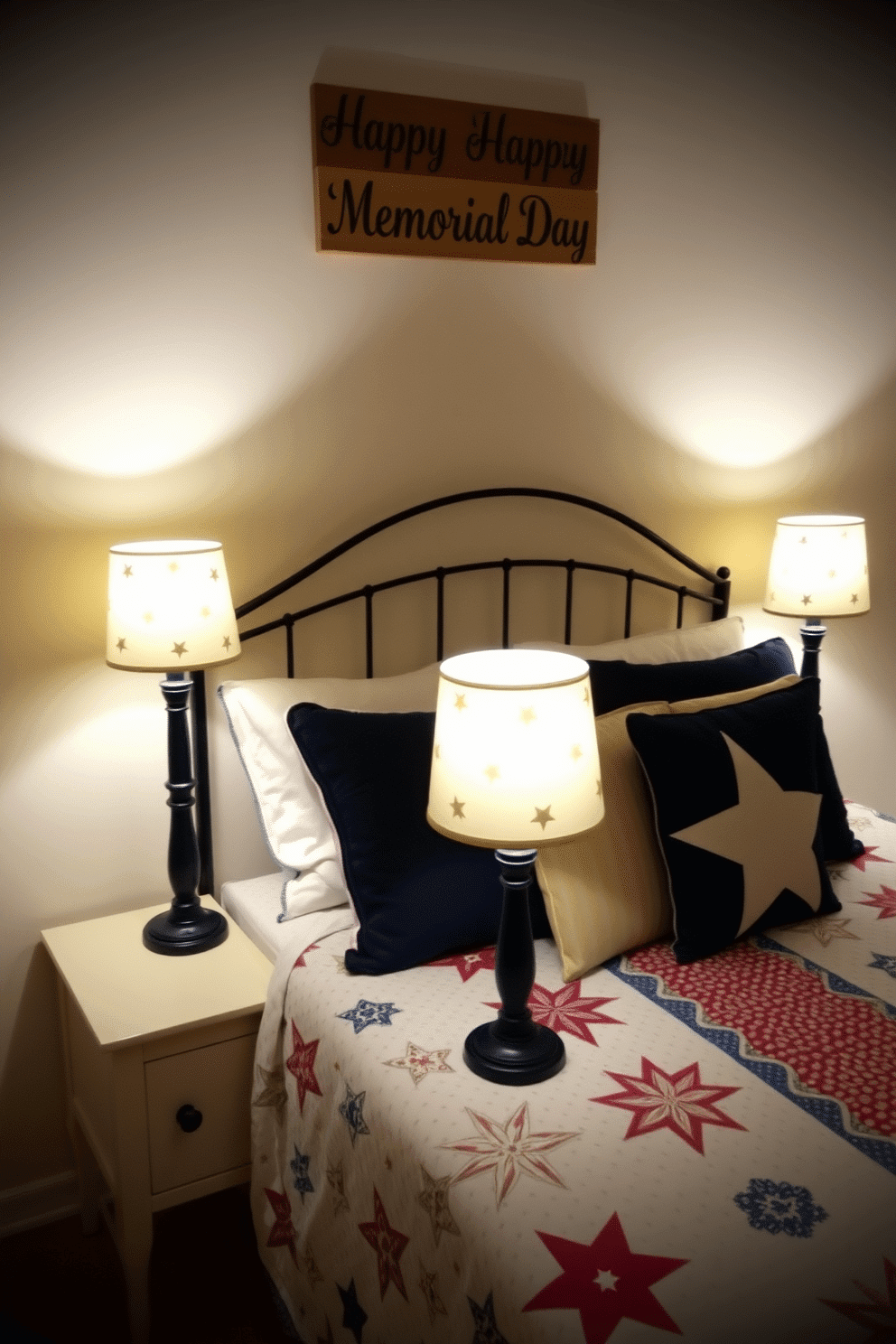 A cozy bedroom setting. Two star-spangled table lamps sit on either side of the bed, each with a navy blue base and a white shade adorned with small, golden stars. The bed is dressed with a quilt featuring red, white, and blue patterns, complemented by matching throw pillows. Above the headboard, a wooden sign with 