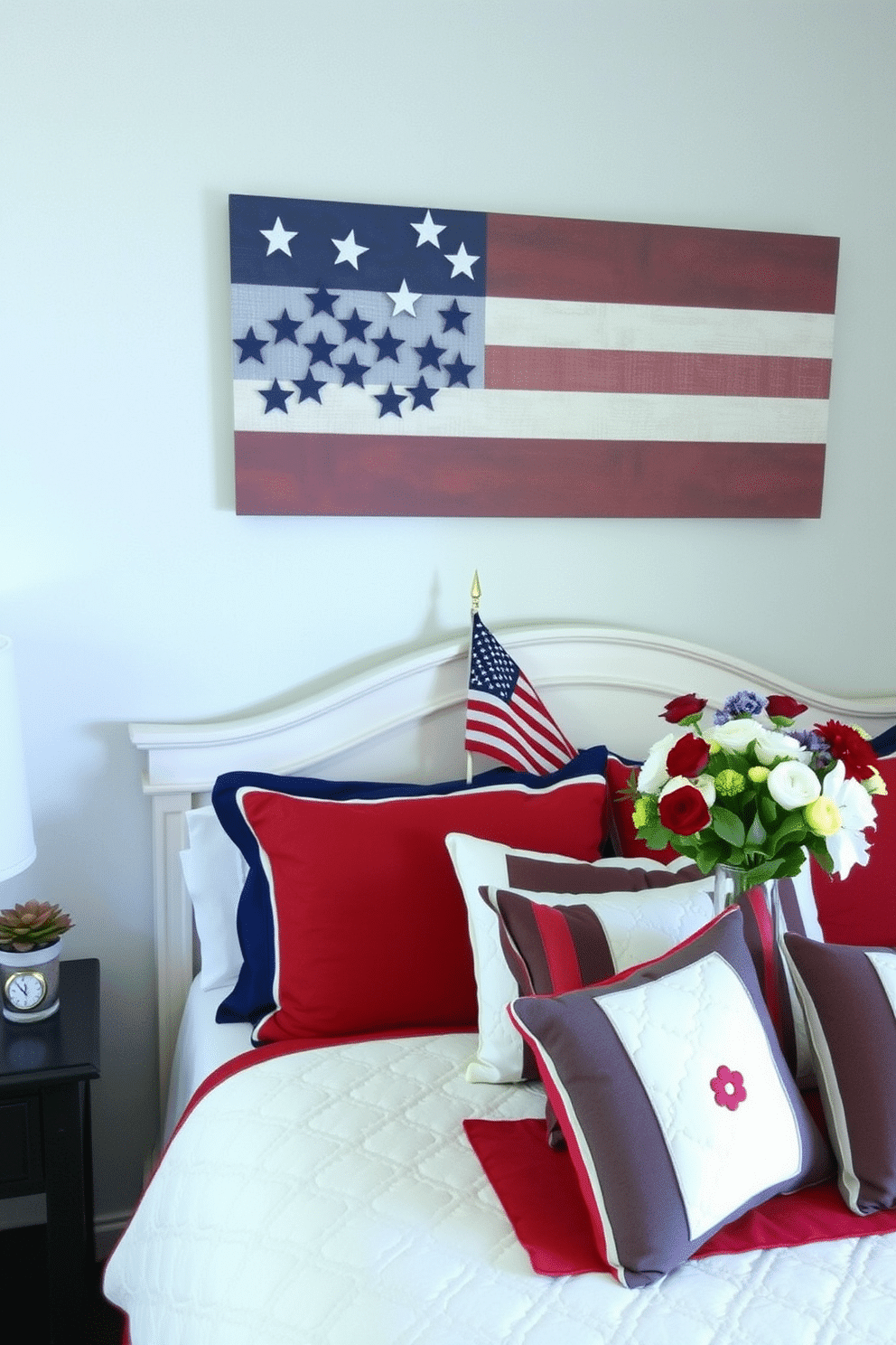 A cozy bedroom decorated for Memorial Day. The walls feature a striking stars and stripes wall art display above the headboard, creating a patriotic focal point. The bed is adorned with red, white, and blue bedding, including a quilted comforter and matching pillows. On the nightstand, there is a small American flag and a vase filled with fresh flowers in patriotic colors.