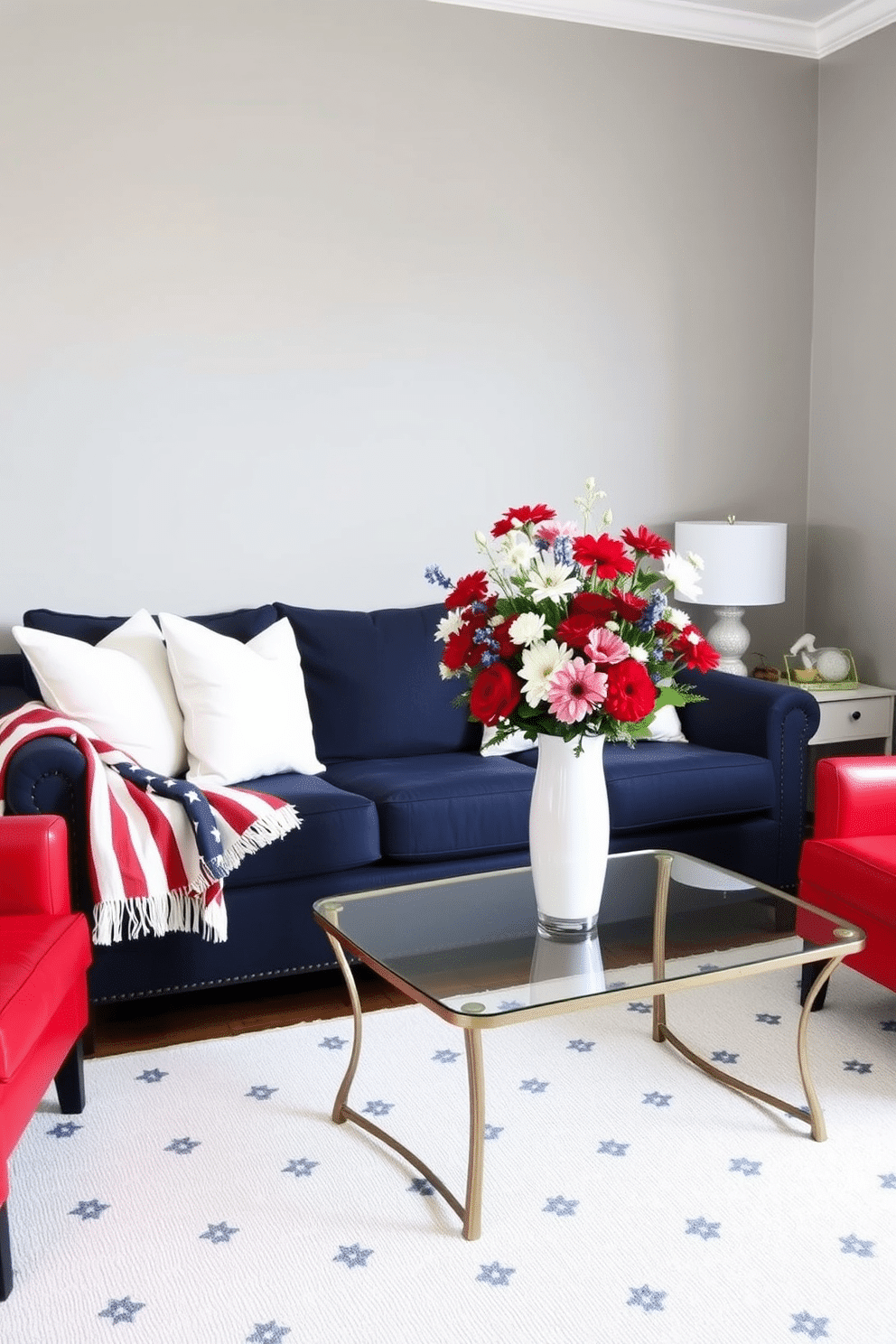 A cozy living room setting. There's a navy blue sofa with white and red throw pillows arranged neatly. In front of the sofa, a coffee table holds a large vase filled with red, white, and blue flowers, creating a festive centerpiece. The walls are painted a soft gray, and an American flag-themed throw blanket is draped over the arm of the sofa. A white rug with subtle blue patterns covers the floor, and a pair of red armchairs flank the coffee table, completing the patriotic look.