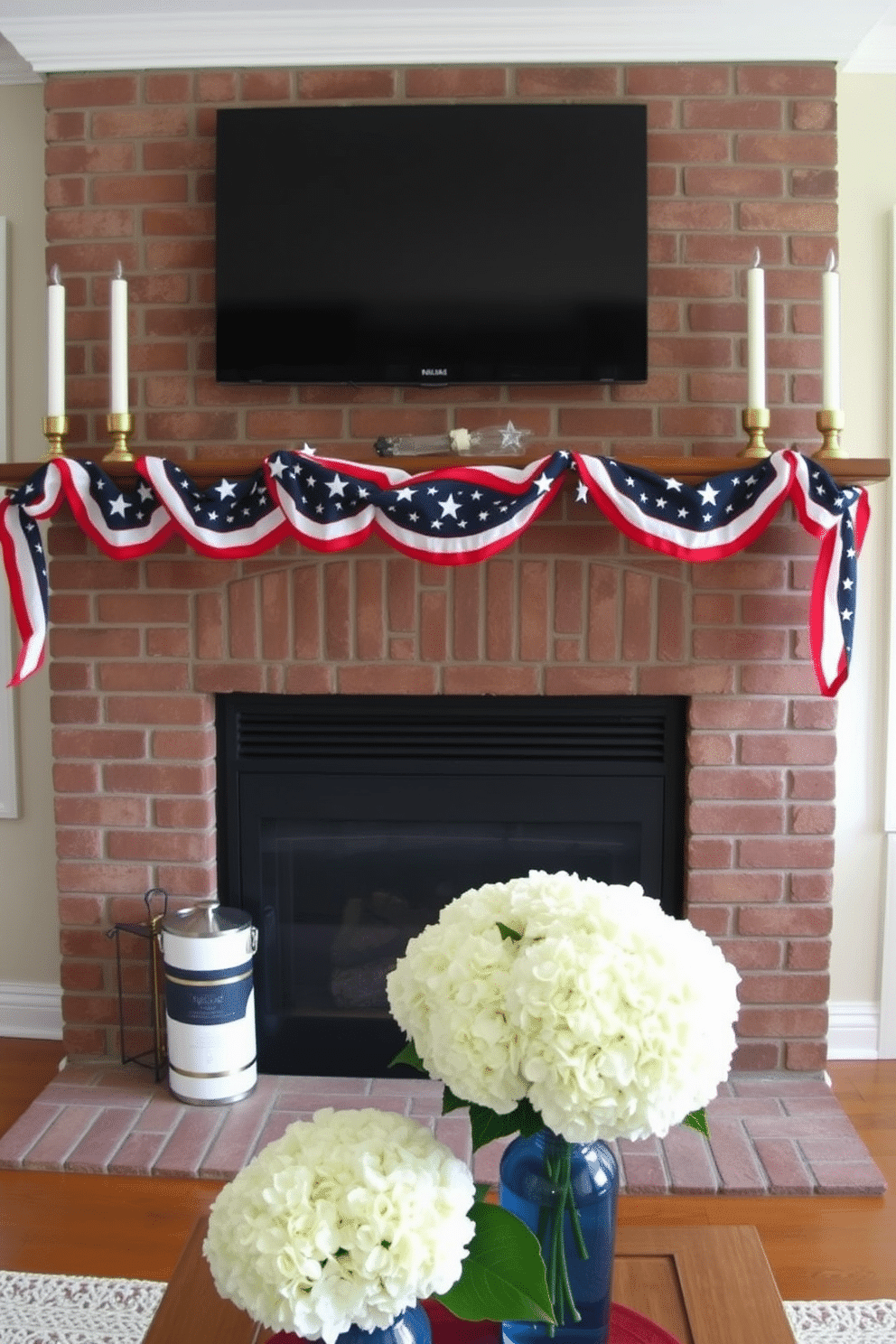 A cozy living room with a classic brick fireplace. The mantel is adorned with a patriotic garland featuring red, white, and blue stars and stripes, draping elegantly across its length. On either side of the mantel, tall, white candles in brass holders add a touch of elegance. Below, a simple yet striking arrangement of white hydrangeas in a blue vase sits on the hearth, completing the festive Memorial Day look.