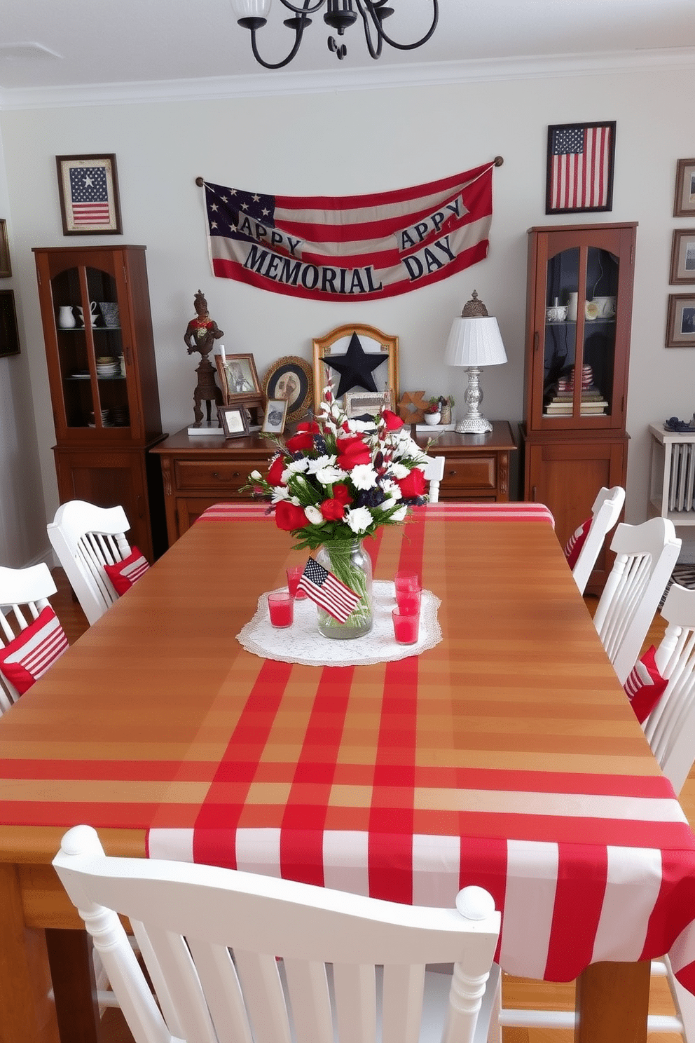 A festive dining room setting. A large wooden dining table is covered with a red and white striped tablecloth, creating a vibrant and patriotic atmosphere. Surrounding the table are white wooden chairs, each adorned with a small American flag pillow. On the table, a centerpiece features a mix of red, white, and blue flowers in a clear glass vase, accompanied by small candles in red holders. The walls are decorated with vintage American flag prints, and a banner reading 