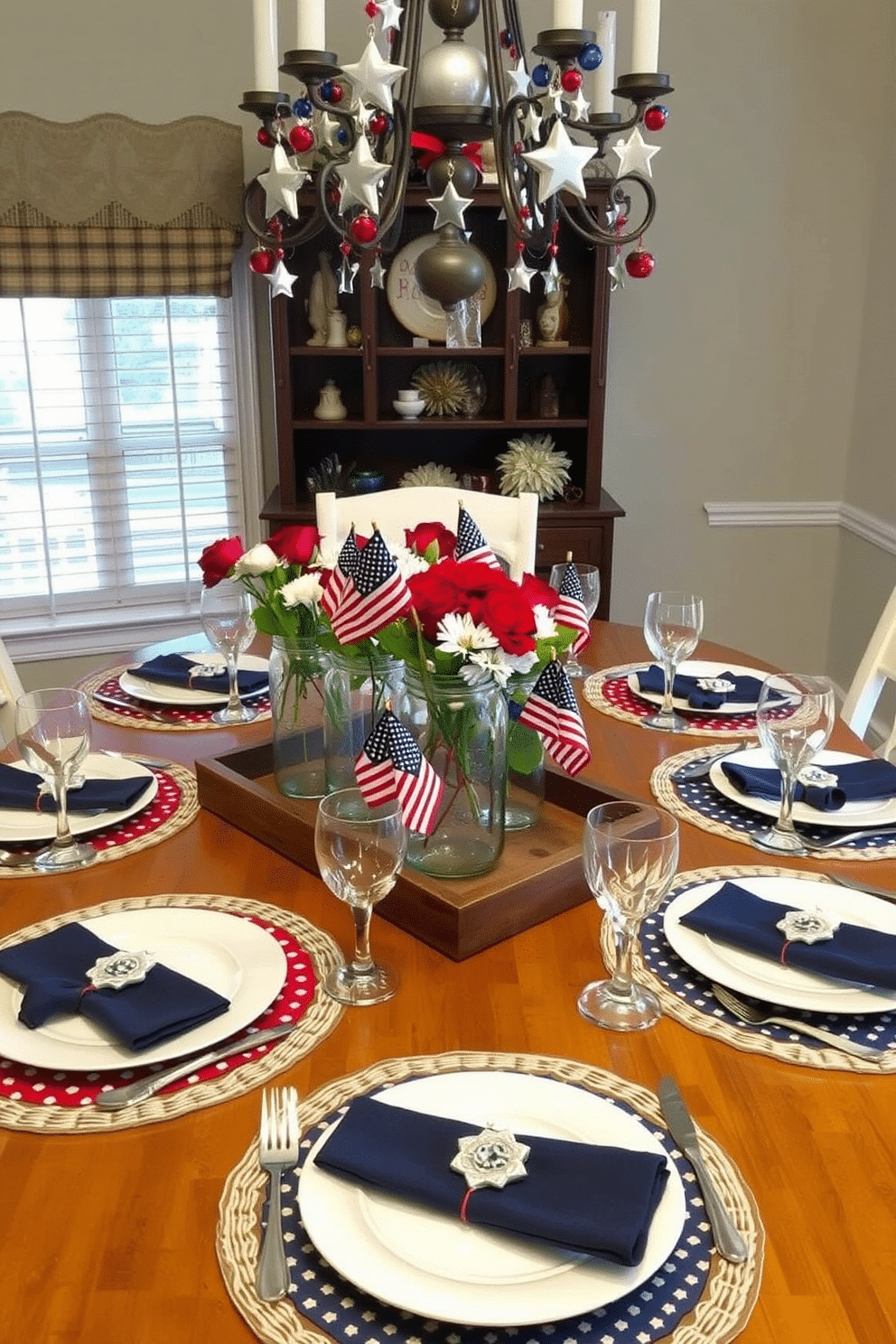 A dining room table set for Memorial Day, featuring layered placemats in patriotic red, white, and blue hues. Each place setting includes a white dinner plate with a blue napkin, topped with a small American flag, and surrounded by silver cutlery and crystal glassware. The centerpiece consists of a rustic wooden tray holding mason jars filled with red, white, and blue flowers. Above the table, a chandelier adorned with miniature American flags and star-shaped ornaments adds a festive touch.