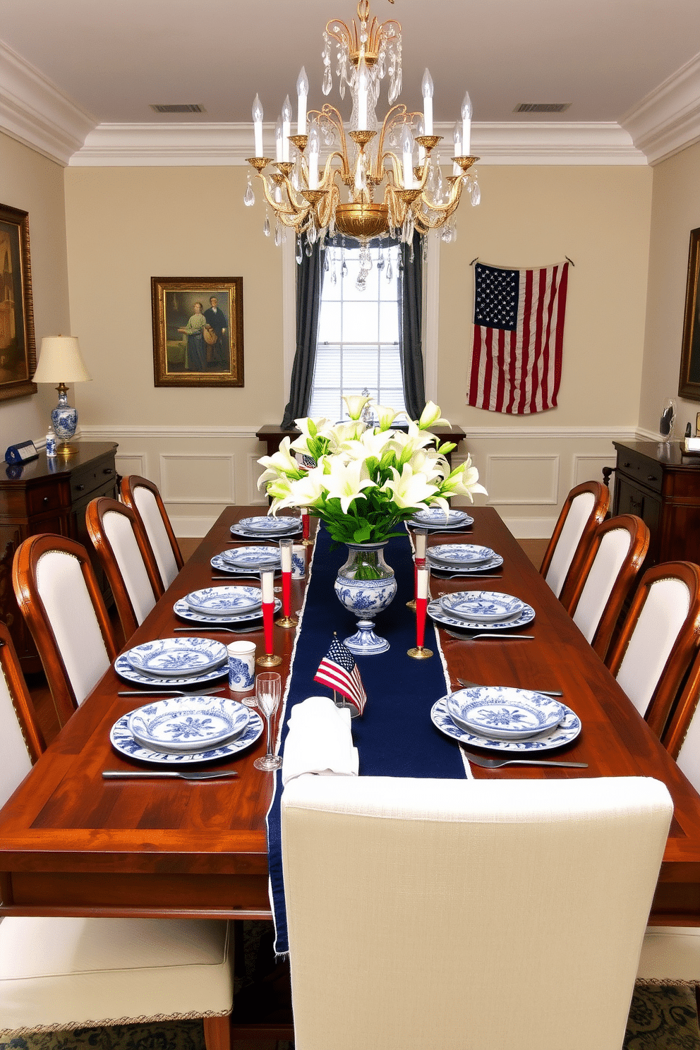 A Memorial Day dining room features a large wooden table set with blue and white porcelain dishware, each piece adorned with intricate floral patterns. The table is surrounded by white upholstered chairs, and a navy blue table runner extends down its length, accented with small American flags and red, white, and blue candles. On the walls, patriotic-themed artwork and vintage American flags add a touch of history and elegance. A crystal chandelier hangs above the table, casting a warm glow over the setting, while a vase of fresh white lilies serves as a centerpiece, completing the festive yet sophisticated ambiance.