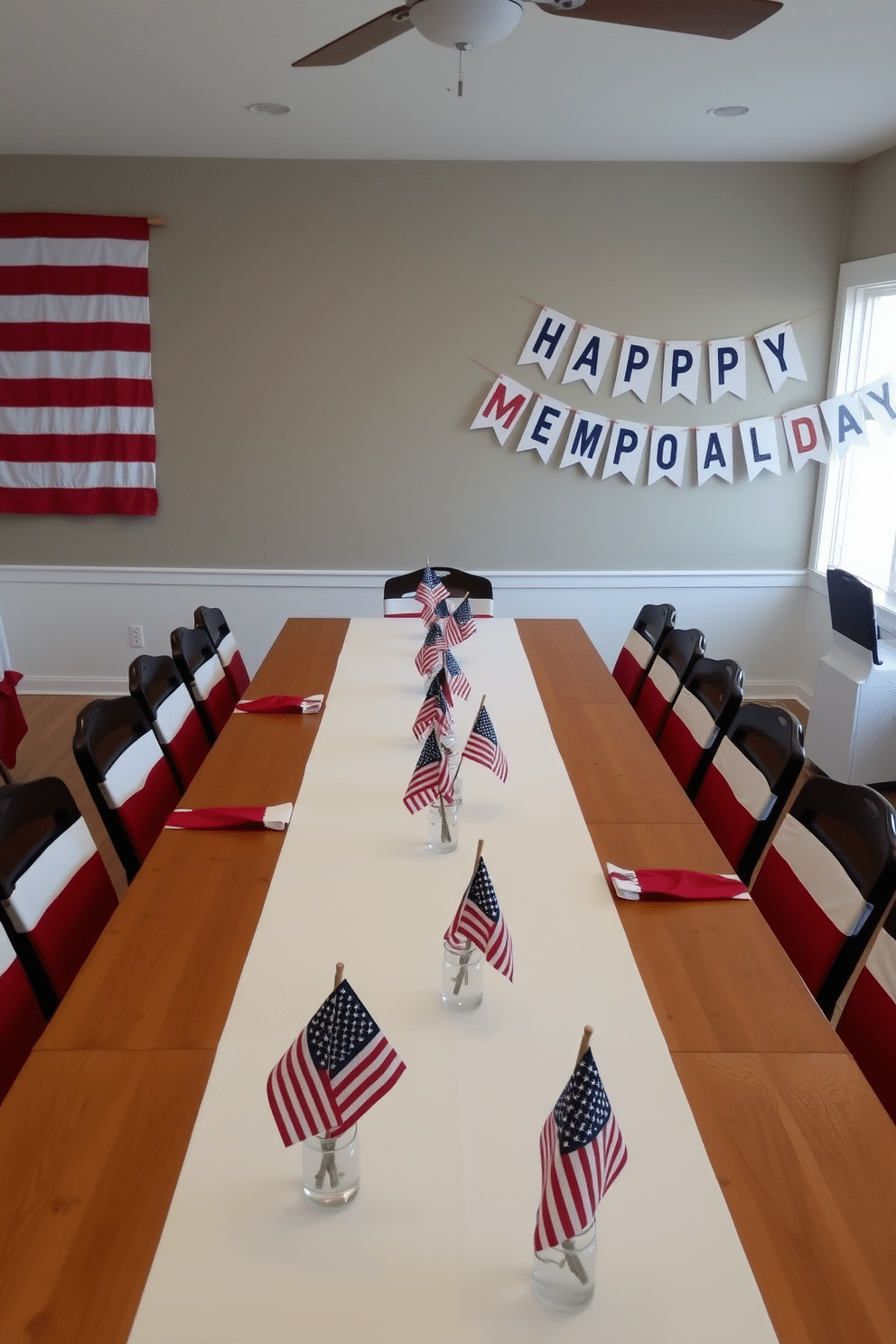 A dining room set for Memorial Day celebrations, featuring a long wooden table adorned with a white tablecloth. Miniature American flags are placed in clear glass vases, evenly spaced along the center of the table, creating a patriotic centerpiece. The room is decorated with red, white, and blue accents, including napkins, plates, and chair covers. A large American flag hangs on one wall, while a festive banner spelling 