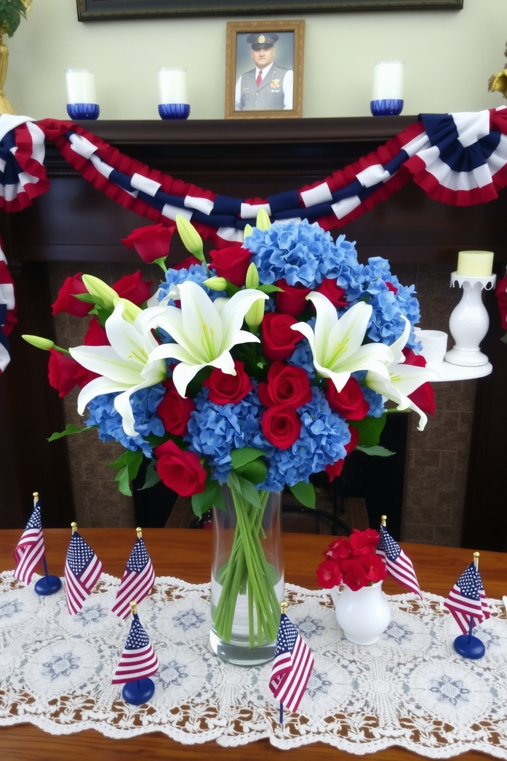 A vibrant floral arrangement featuring red roses, white lilies, and blue hydrangeas displayed in a clear glass vase. The vase is placed on a wooden table with a white lace tablecloth, surrounded by small American flags. A Memorial Day mantel decorated with red, white, and blue garlands draped across the front. On the mantel, there are white candles in blue holders, a framed photo of a veteran, and a small bouquet of red poppies in a white ceramic vase.