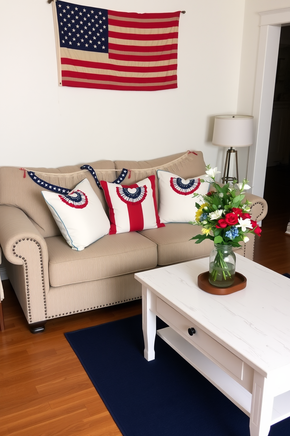 A small living room decorated for Memorial Day. Mini flag banners are draped across the back of a cozy beige sofa and along the edge of a white wooden coffee table. Red, white, and blue throw pillows are placed on the sofa, complementing a navy blue area rug. A vintage American flag hangs on the wall above the sofa, and a vase with fresh flowers in patriotic colors sits on the coffee table.