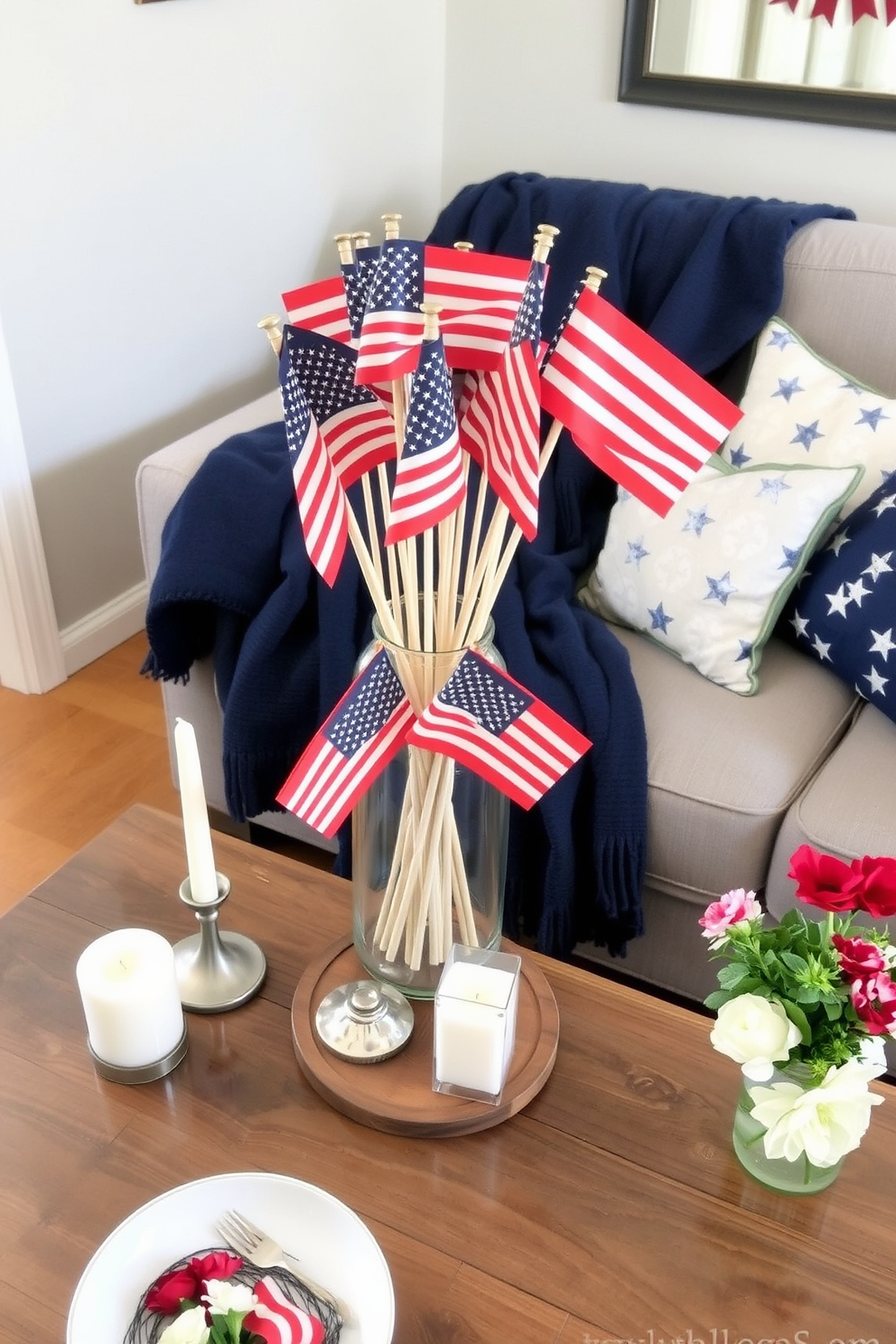 A charming centerpiece featuring a collection of miniature flags arranged in a clear vase. The vase sits on a rustic wooden table, surrounded by simple white candles and fresh flowers for a festive touch. The small space is decorated with red, white, and blue accents to celebrate Memorial Day. A cozy seating area includes a navy blue throw blanket draped over a light gray couch, complemented by star-patterned pillows.