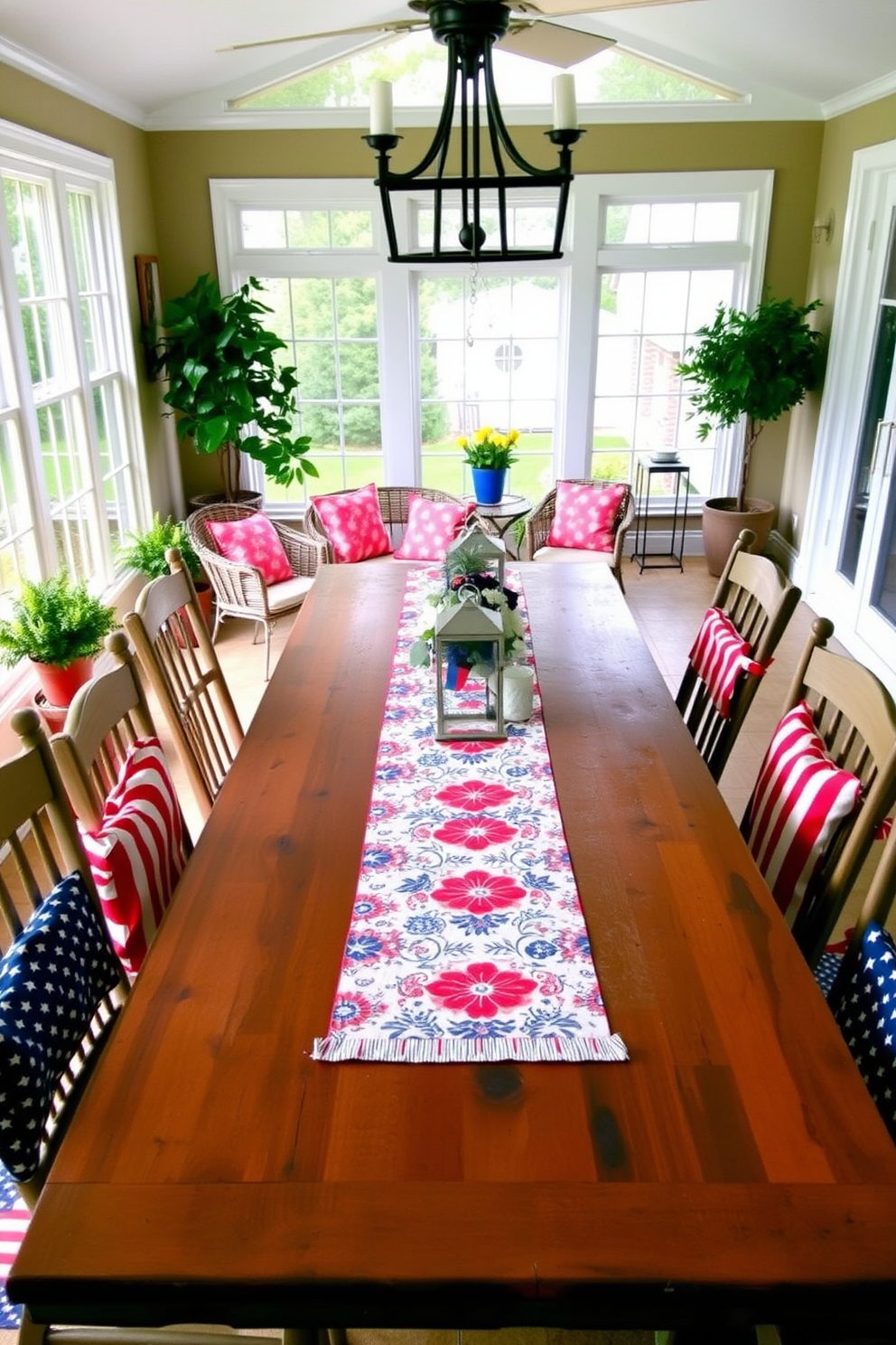 A festive table runner adorns a long wooden dining table, featuring vibrant red, white, and blue patterns that celebrate Memorial Day. Surrounding the table are mismatched chairs, each draped with a coordinating fabric that enhances the cheerful atmosphere. In the sunroom, large windows allow natural light to flood the space, highlighting a cozy seating area with plush cushions in patriotic colors. Potted plants and decorative lanterns are strategically placed to create a warm and inviting ambiance for gatherings.