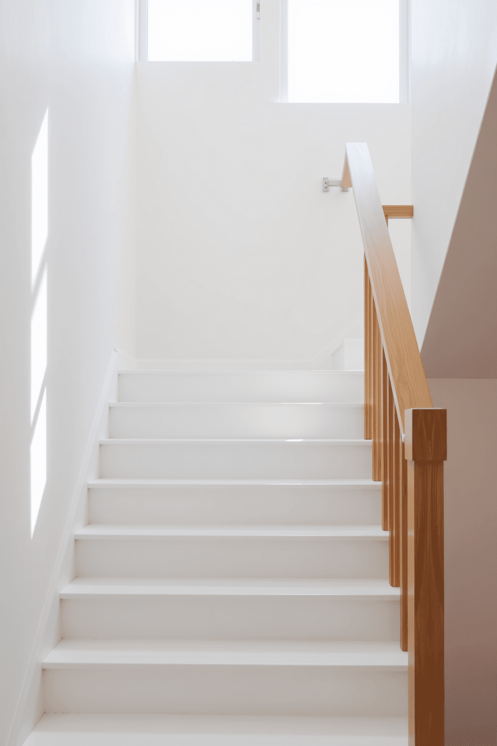 A clean and airy staircase featuring simple white steps complemented by natural wood accents. The handrail is crafted from polished wood, seamlessly blending with the minimalist aesthetic of the space. Natural light floods the area through a large window, casting soft shadows on the stairs. The surrounding walls are painted in a light neutral tone, enhancing the staircase's elegance and simplicity.