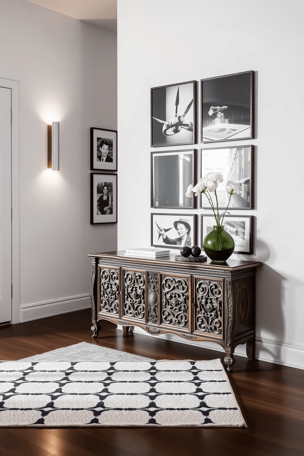 A modern entryway featuring a vintage console table with intricate carvings, paired with sleek contemporary wall-mounted lighting. The walls are adorned with a gallery of framed black-and-white photographs, and a bold geometric rug anchors the space.