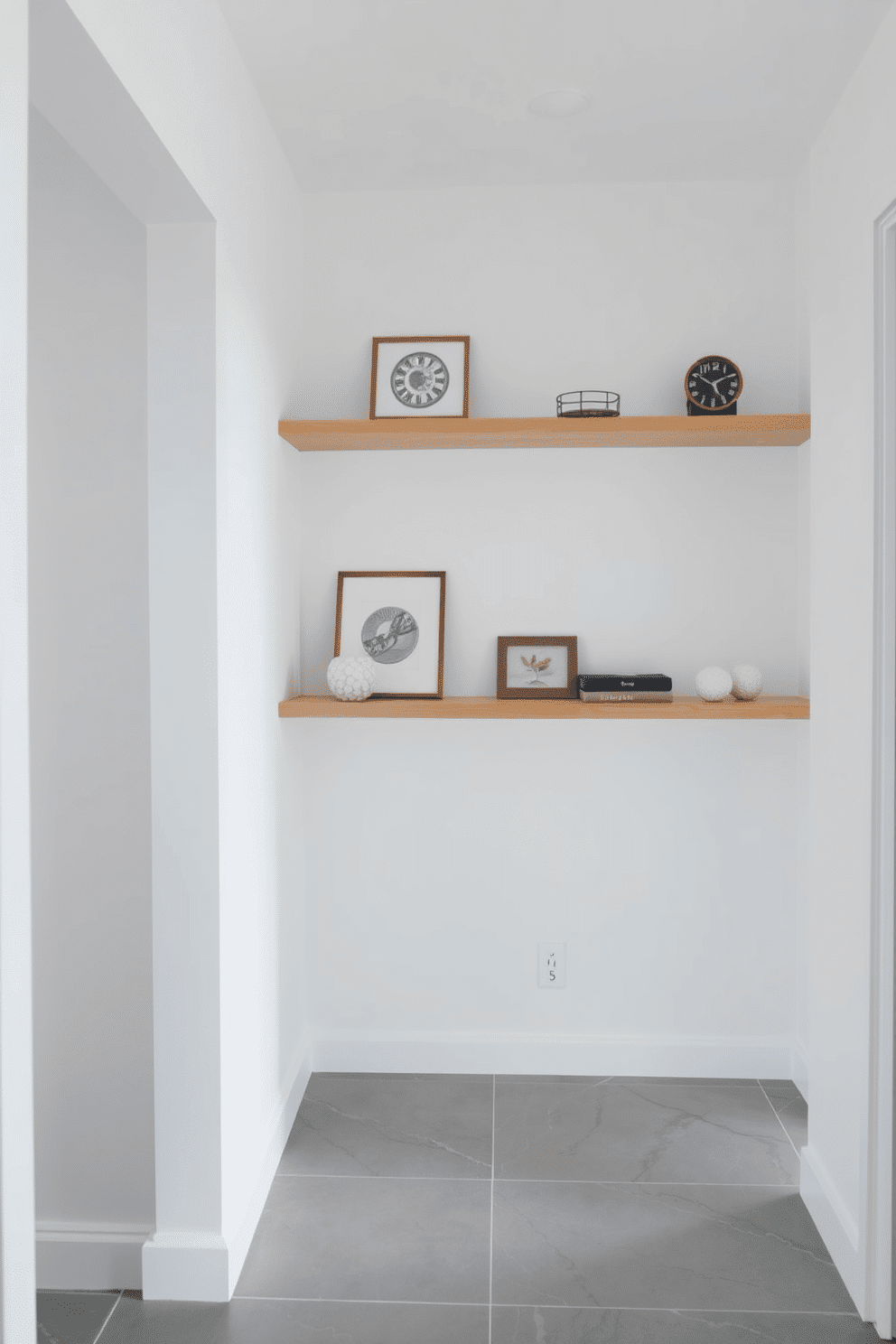 A modern entryway featuring floating shelves adorned with carefully curated decorative items. The walls are painted in a crisp white, while the floor showcases a sleek gray tile, creating a bright and inviting atmosphere.