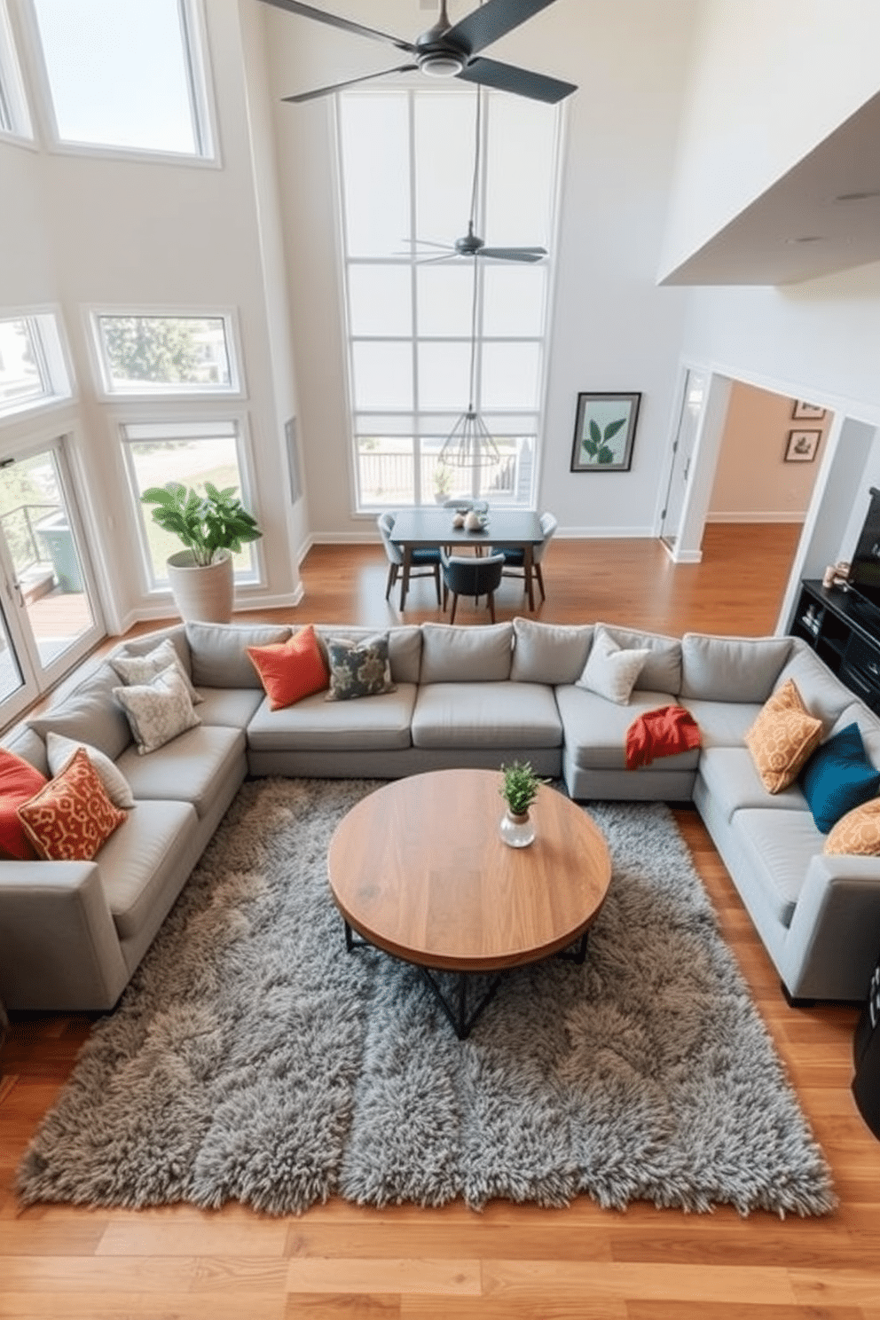 Open concept layout with cozy seating. The family room features a large sectional sofa in soft gray, accented by colorful throw pillows. A round wooden coffee table sits in the center, surrounded by plush area rugs that add warmth to the space. Large windows allow natural light to flood in, highlighting the minimalist decor and a few carefully chosen art pieces on the walls.