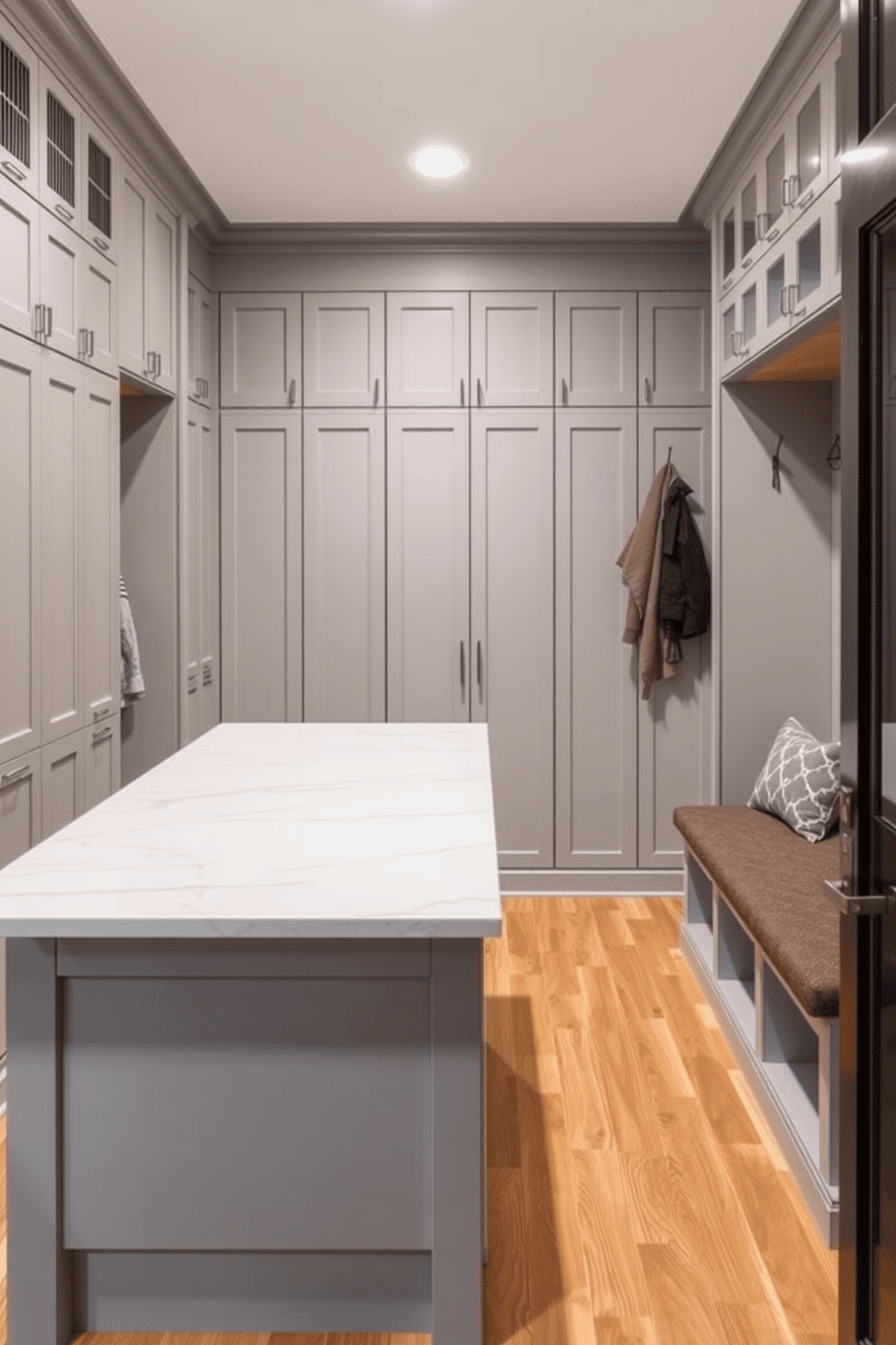 A modern mudroom featuring a spacious island designed for extra workspace. The island is topped with a sleek quartz surface and surrounded by built-in storage cabinets painted in a soft gray hue. On one side, there are hooks for coats and a bench upholstered in a durable fabric for seating. The flooring is a stylish, water-resistant vinyl that mimics natural wood, adding warmth to the space.