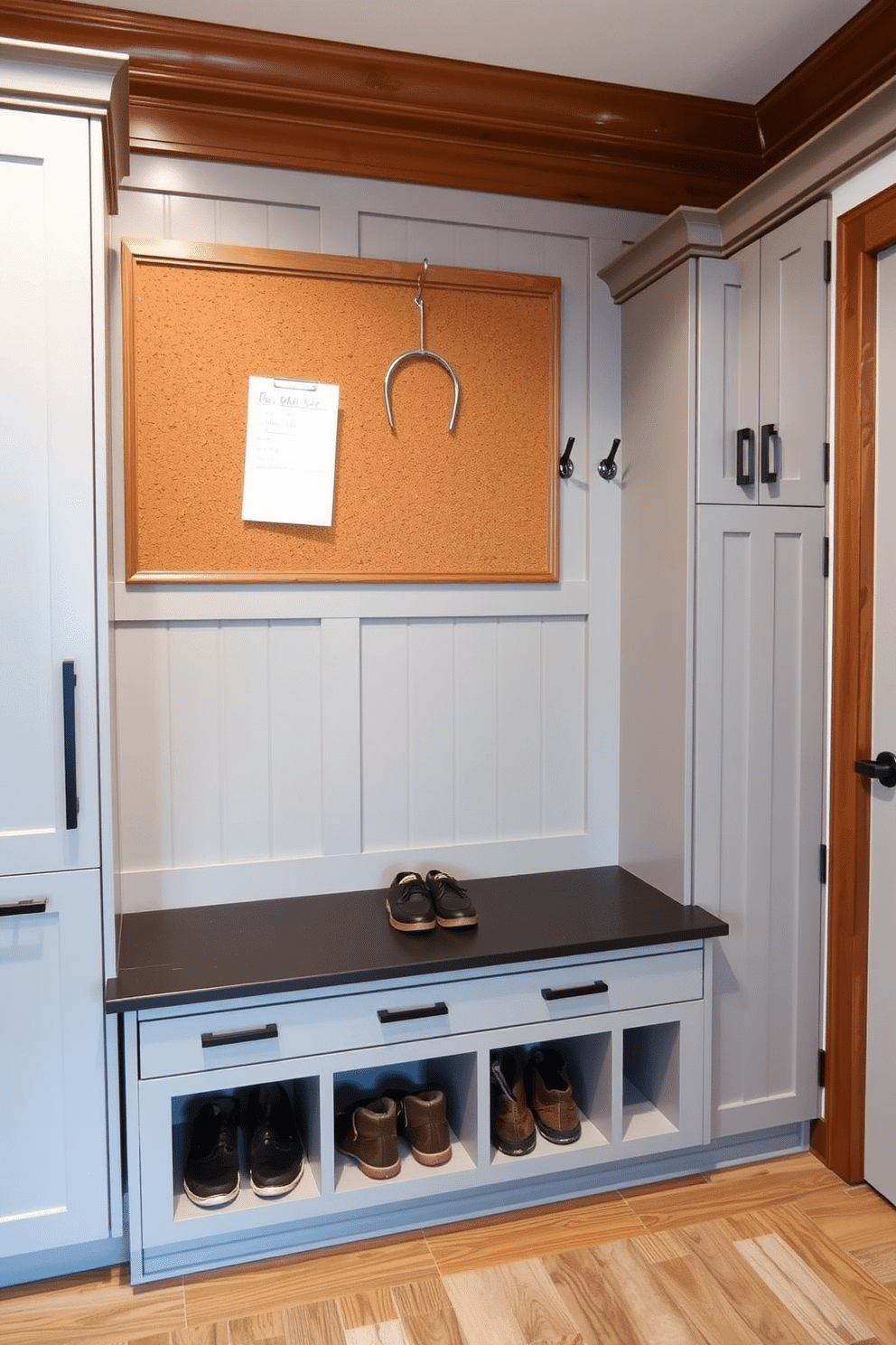 A functional corkboard wall serves as an organized space for notes and reminders, framed by sleek wooden trim. Below, a built-in bench with storage cubbies provides a practical seating area for removing shoes, while hooks above hold coats and bags. The modern mudroom features a combination of light gray cabinetry and contrasting dark hardware, creating a stylish yet practical entryway. A durable tile floor mimics the look of hardwood, ensuring easy maintenance and a warm welcome.