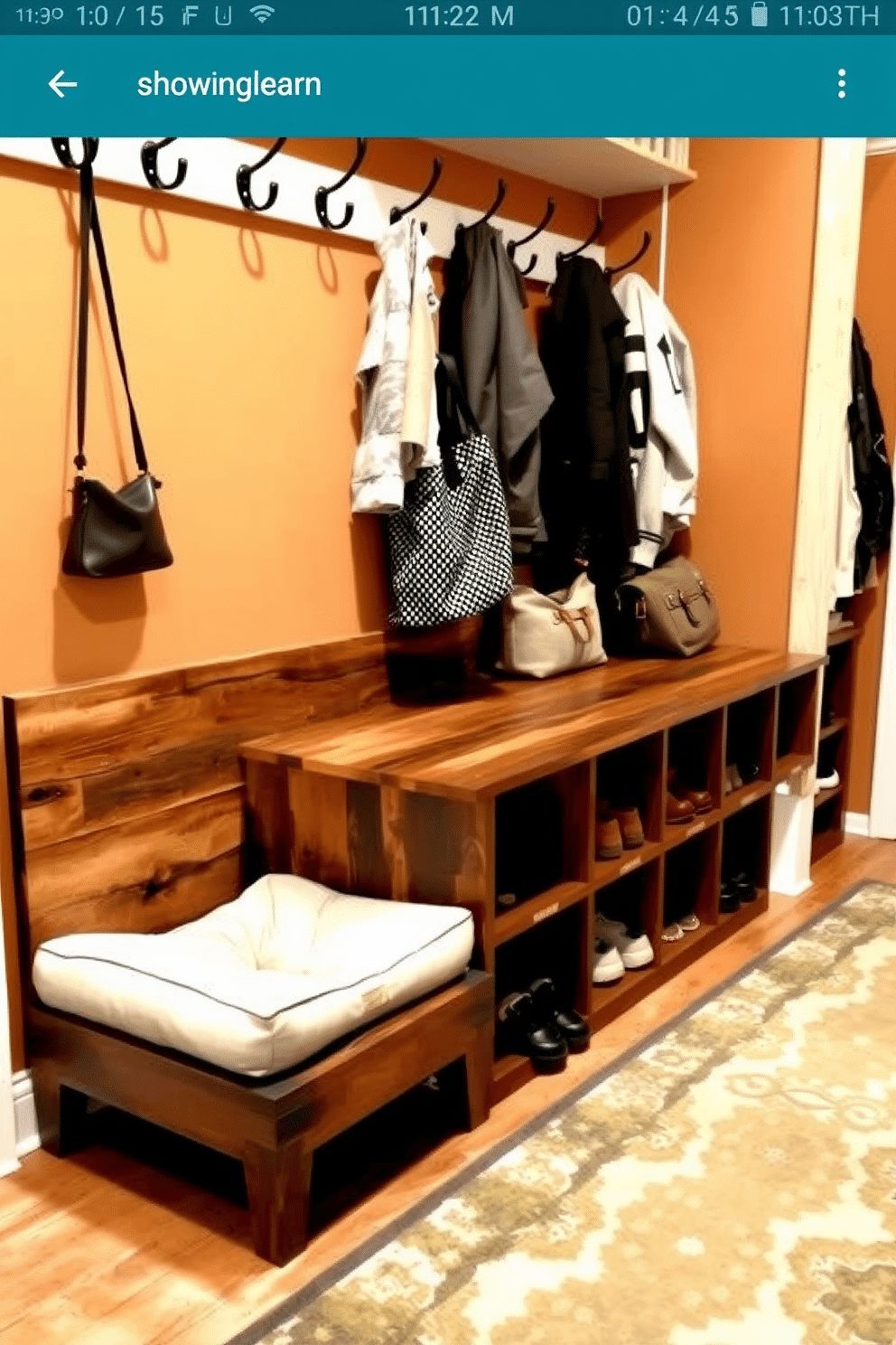 A stylish mudroom features a bench with an integrated pet bed underneath, combining functionality with comfort. The bench is crafted from reclaimed wood, while the pet bed is cushioned with soft, machine-washable fabric in a neutral tone to complement the space. Above the bench, hooks are mounted for coats and bags, and a row of cubbies provides storage for shoes and outdoor gear. The walls are painted in a warm, inviting color, and a patterned rug adds a touch of personality to the entryway.