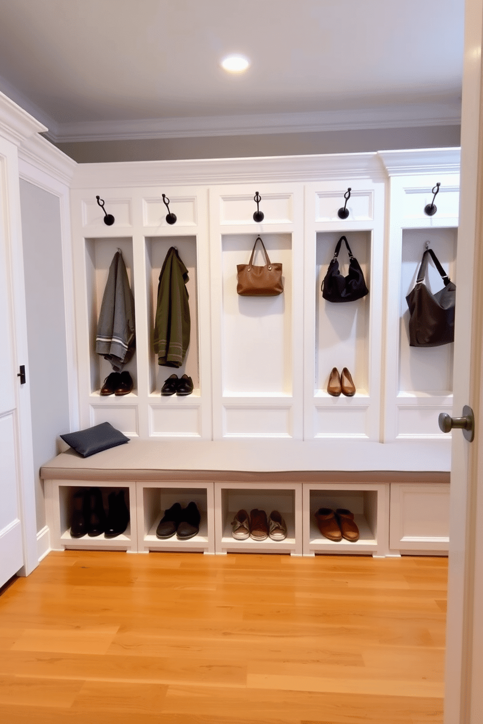 Elegant white cubbies with crown molding are arranged neatly along the wall, providing ample storage for shoes and outdoor gear. The space is accented with a soft gray bench, upholstered in a durable fabric, inviting guests to sit while they remove their shoes. Above the cubbies, decorative hooks are installed for hanging coats and bags, creating a functional yet stylish entryway. The floor is finished with a warm wood tone, complementing the crisp white cabinetry and enhancing the overall inviting atmosphere of the mudroom.