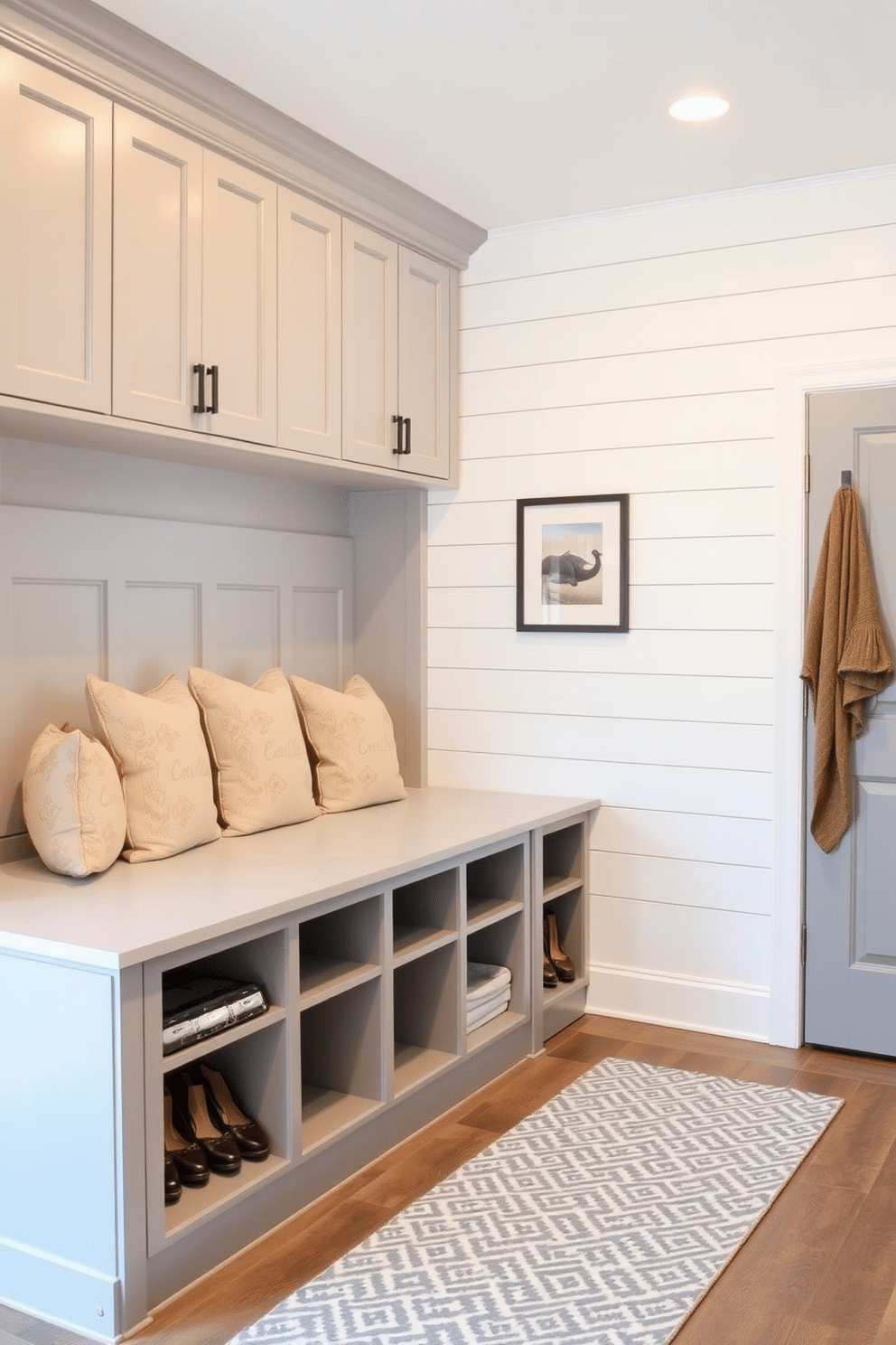 A stylish mudroom island features built-in seating with plush cushions and ample storage beneath. The island is framed by cabinetry in a soft gray finish, while the walls are adorned with shiplap in a crisp white, creating a bright and welcoming atmosphere.