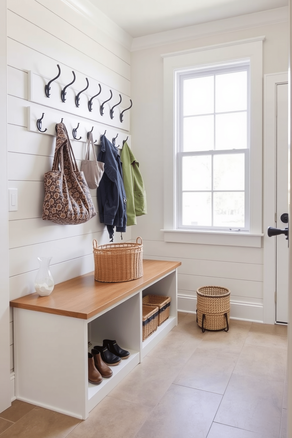 A functional bench with storage underneath is positioned against the wall, offering a practical solution for organizing shoes and outdoor gear. Above the bench, a series of hooks are mounted for coats and bags, while a large window allows natural light to brighten the space. The mudroom features a combination of durable materials, including a tile floor that is easy to clean and a shiplap accent wall for added texture. A stylish basket sits on the bench, providing a decorative yet functional element to this inviting laundry room design.