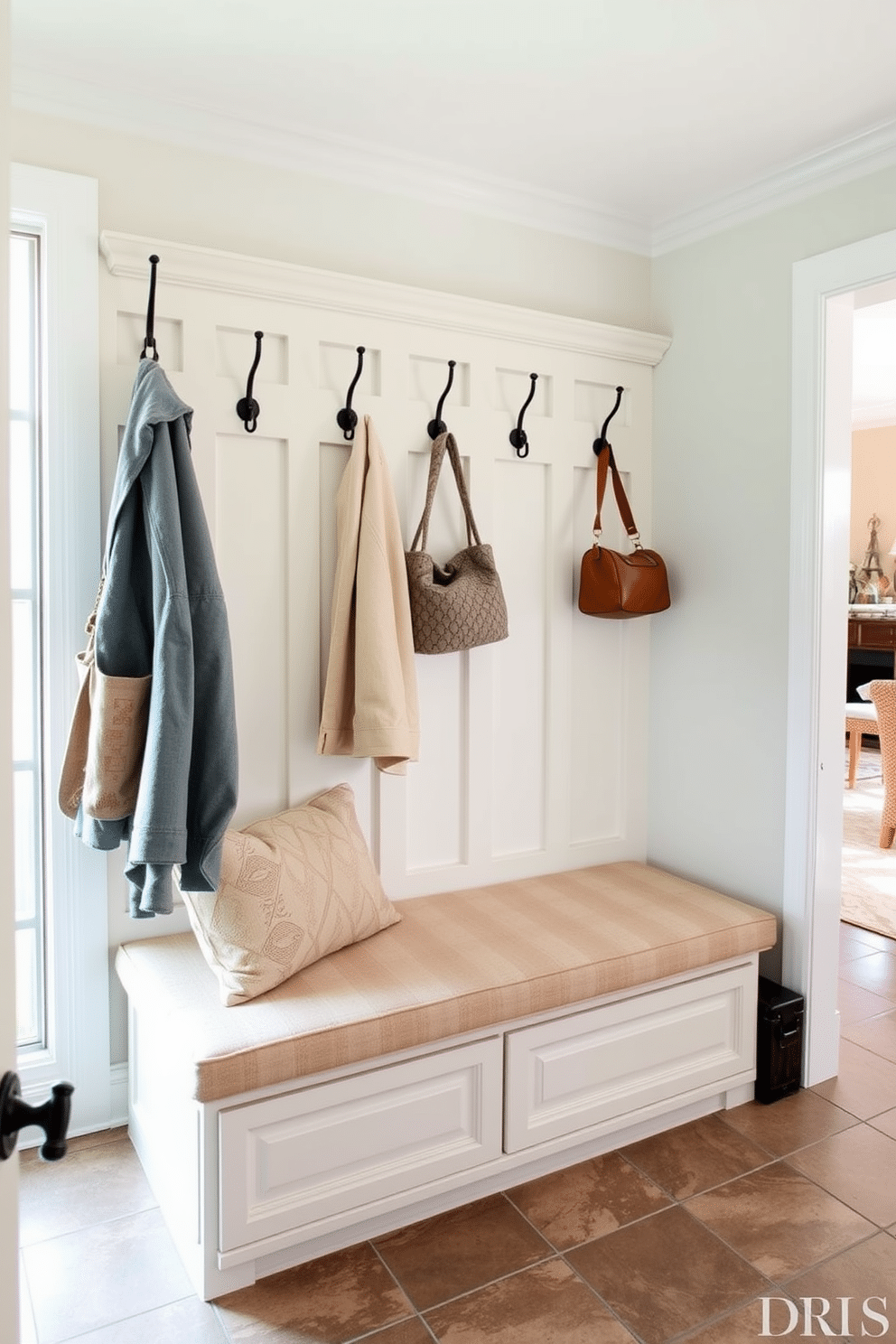 A cozy bench with storage underneath is positioned against the wall, upholstered in soft, durable fabric that complements the room's color palette. Above the bench, hooks are mounted for hanging coats and bags, while a decorative cushion adds a touch of comfort. The mudroom features tiled flooring for easy cleaning, seamlessly connecting to the adjacent family room. Natural light floods the space through a large window, highlighting the organized storage solutions and creating an inviting atmosphere.