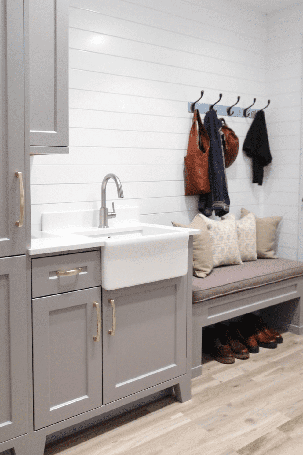 A functional mudroom sink is designed with a deep basin, perfect for washing off muddy shoes and hands. The cabinetry is painted in a soft gray, complemented by a white quartz countertop that features a stylish, brushed nickel faucet. Adjacent to the sink, a built-in bench with plush cushions provides a cozy spot for putting on or taking off shoes. The walls are adorned with shiplap, and hooks for coats and bags are neatly arranged above the bench, enhancing the room's practicality and charm.