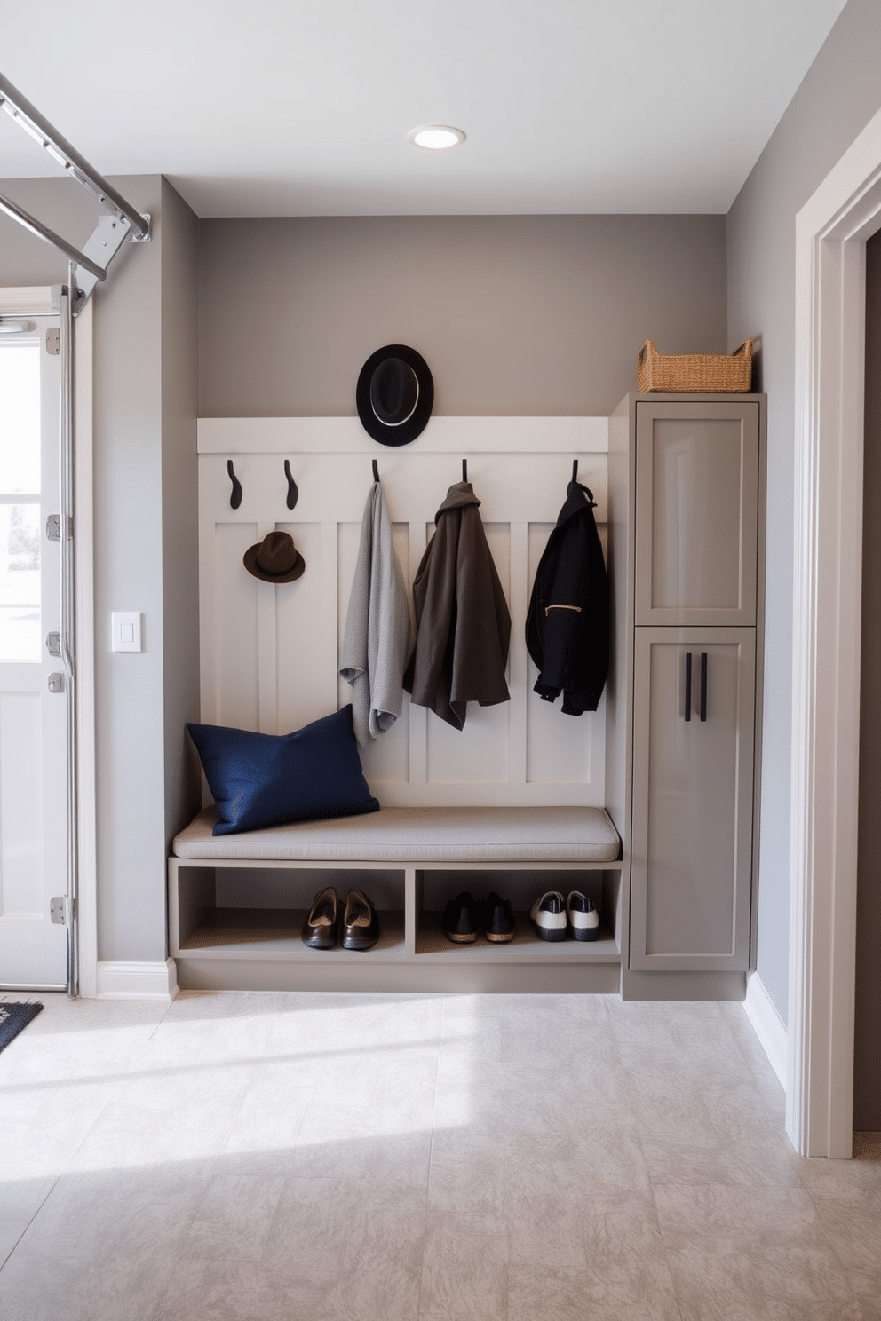 A stylish mudroom near the garage features a wall-mounted bench with a cushioned seat, providing comfort while putting on shoes. Hooks for coats and hats are neatly arranged above the bench, complemented by a sleek storage cabinet for shoes and outdoor gear. The walls are painted in a soft gray hue, creating a calm and inviting atmosphere. Natural light floods the space through a nearby window, highlighting the textured tile flooring that adds durability and style.