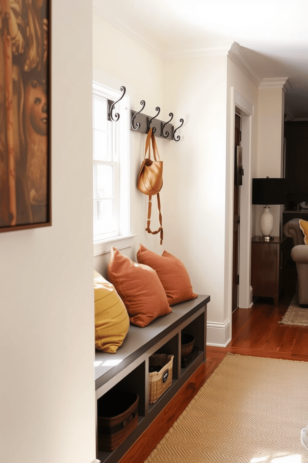 A cozy bench with storage underneath is positioned against the wall, featuring soft cushions in warm, earthy tones. The space is illuminated by natural light streaming through a nearby window, enhancing the welcoming atmosphere. The mudroom flows seamlessly into the living room, showcasing a harmonious blend of functional design and aesthetic appeal. Decorative hooks line the wall above the bench, providing a practical yet stylish solution for hanging coats and bags.