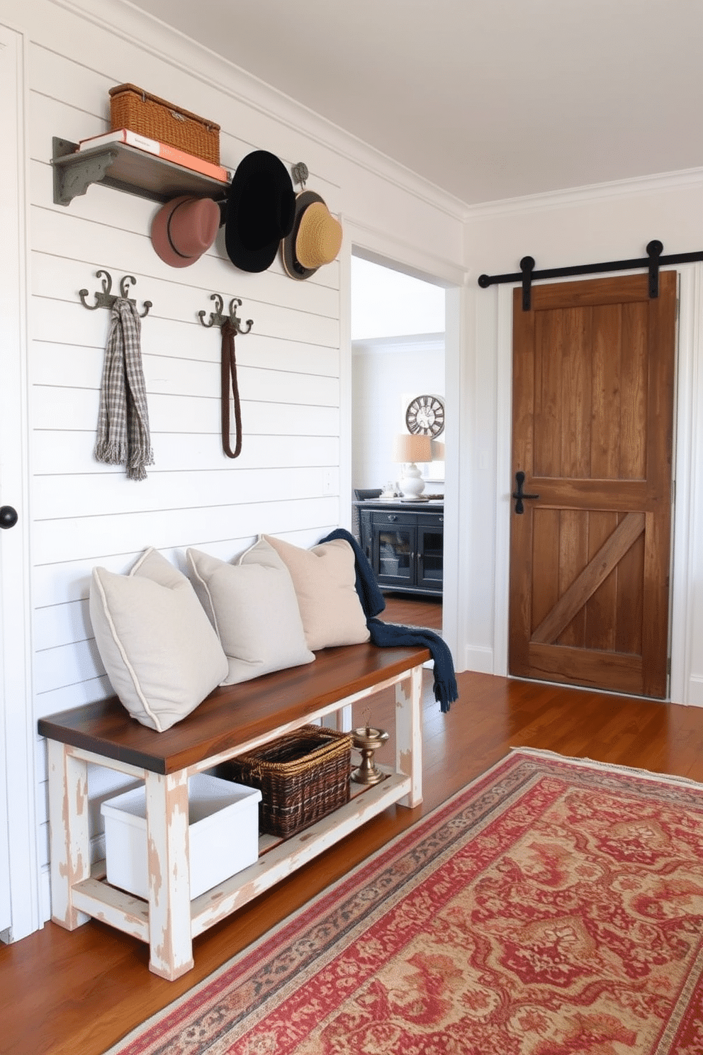 A cozy mudroom designed with vintage accents features a distressed wooden bench adorned with plush cushions. The walls are lined with shiplap painted in a soft, creamy white, while antique hooks hold weathered coats and hats. A rustic sliding barn door separates the mudroom from the living room, adding character and charm. The floor is covered with a patterned vintage rug that complements the room's warm color palette.