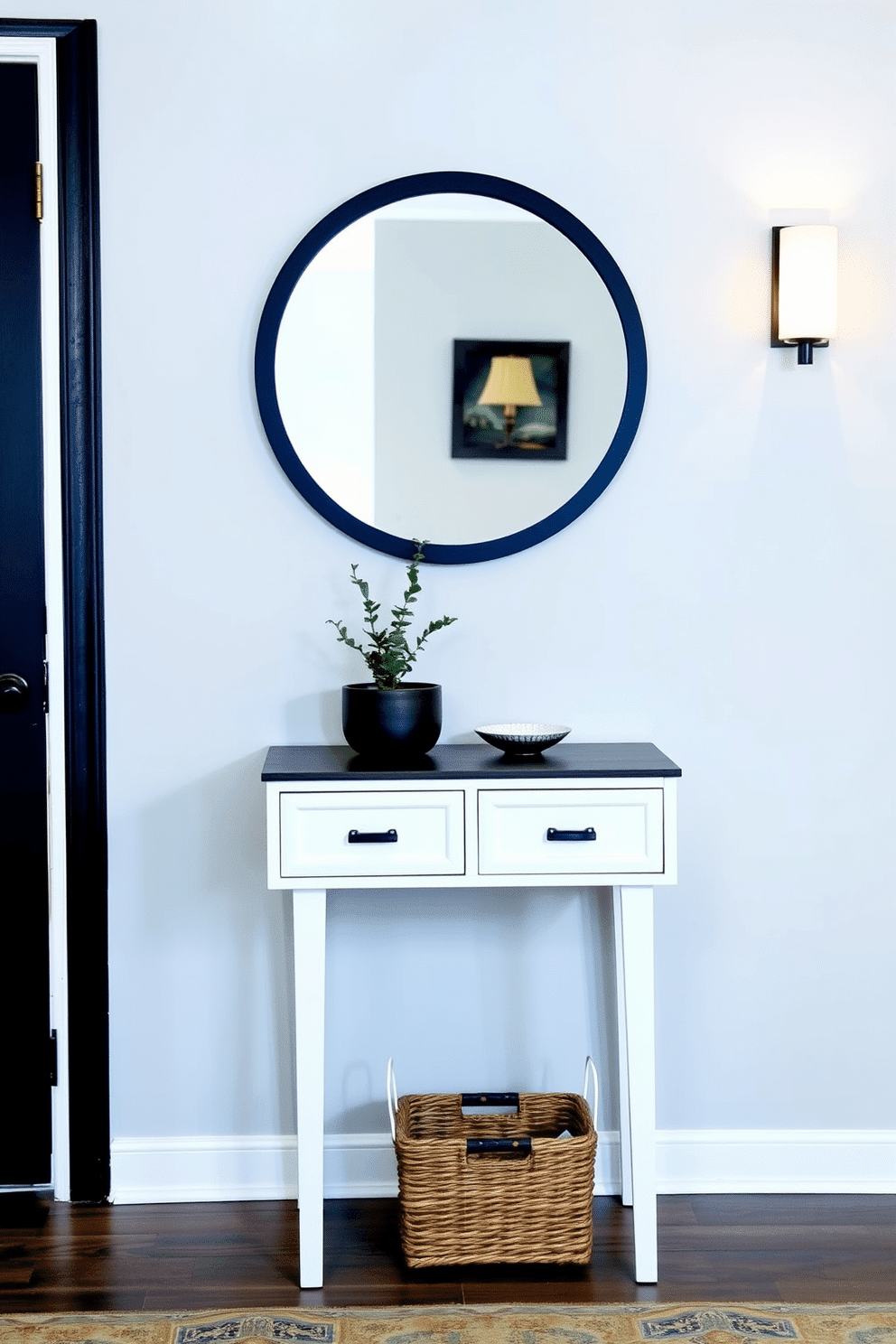 A compact entryway table with drawers stands against a light gray wall, offering a functional yet stylish storage solution. Above the table, a round mirror with a black frame reflects the soft glow of a nearby wall sconce, enhancing the welcoming atmosphere. The table is adorned with a small potted plant and a decorative bowl, adding a touch of greenery and personality. Beneath, a woven basket neatly holds shoes, maintaining an organized and inviting entryway space.