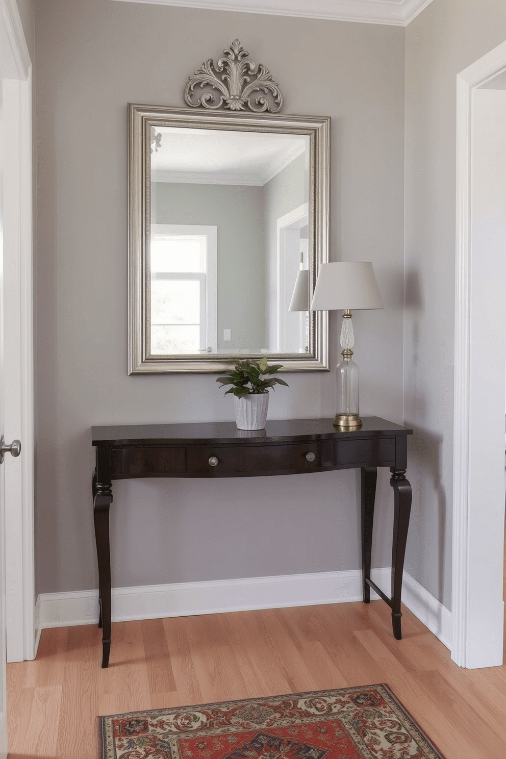 An elegant console table is positioned against a soft gray wall, reflecting natural light from a nearby window. Above the table, a large, ornate wall mirror with a silver frame enhances the sense of space and sophistication. The narrow foyer features a muted color palette with light hardwood flooring, creating a warm and inviting atmosphere. A stylish runner rug adds texture, while a small potted plant sits gracefully on the console table, bringing a touch of greenery to the design.