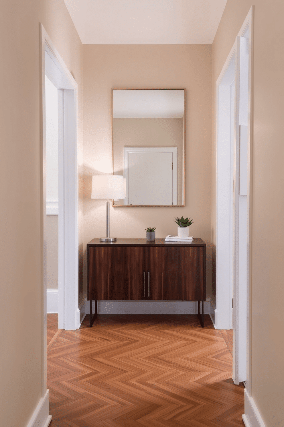 A narrow foyer with a cohesive color palette that creates a sense of harmony. The walls are painted in soft beige, complemented by a sleek console table in a deep walnut finish, adorned with a minimalist lamp and a small potted plant. The flooring features herringbone-patterned hardwood that adds warmth and texture. A large mirror with a thin gold frame hangs above the console, reflecting light and enhancing the space's openness.