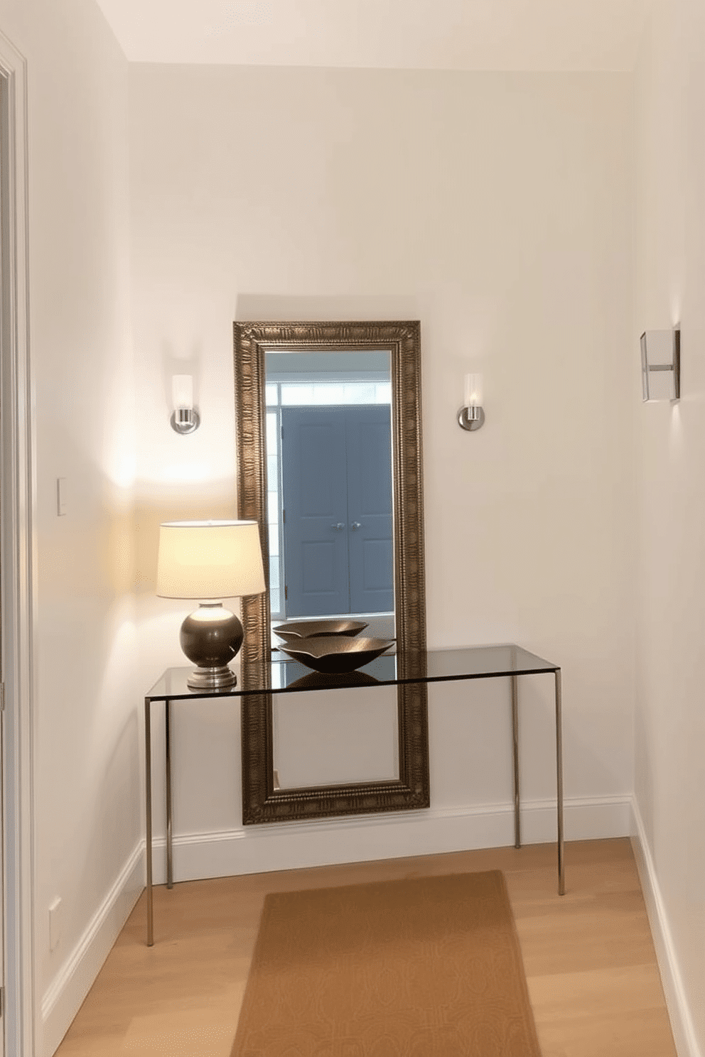 A narrow foyer features a sleek console table against one wall, topped with a stylish lamp and a decorative bowl. Opposite the table, a large mirror with an ornate frame reflects light, creating an illusion of a more spacious entryway. The walls are painted in a light, neutral color to enhance brightness, while a runner rug adds warmth and texture. Flanking the mirror, small wall sconces provide additional illumination, making the space feel inviting and open.