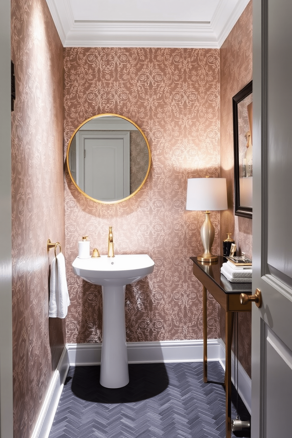 A narrow powder room featuring wallpaper with metallic accents that shimmer softly in the light. The space is adorned with a sleek pedestal sink and a round mirror framed in brushed gold, creating an elegant focal point. On the opposite wall, a small console table holds decorative items and a stylish lamp, enhancing the room's ambiance. The floor is covered with dark herringbone tiles, adding depth and contrast to the overall design.