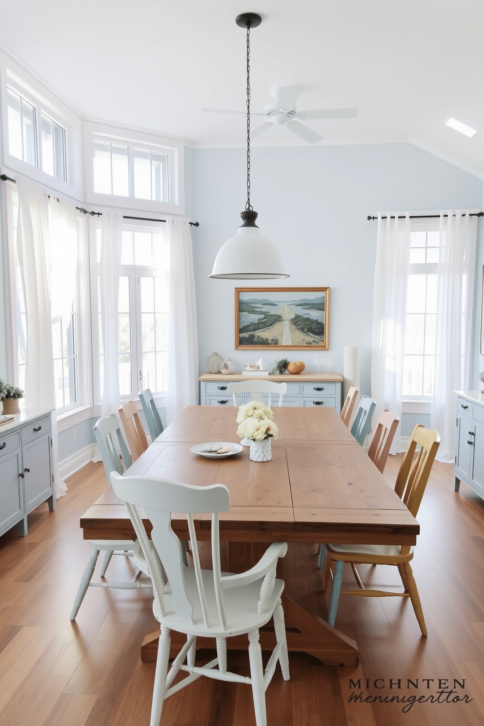 A bright and airy coastal dining space features large windows that invite natural light, adorned with sheer white curtains that flutter gently in the breeze. The room is painted in soft blues and whites, complementing a large wooden dining table surrounded by mismatched chairs in pastel tones. In the center, a statement pendant light hangs above the table, casting a warm glow during evening meals. Coastal-inspired decor, including seashell centerpieces and nautical-themed artwork, enhances the relaxed atmosphere of this open floor plan dining room.