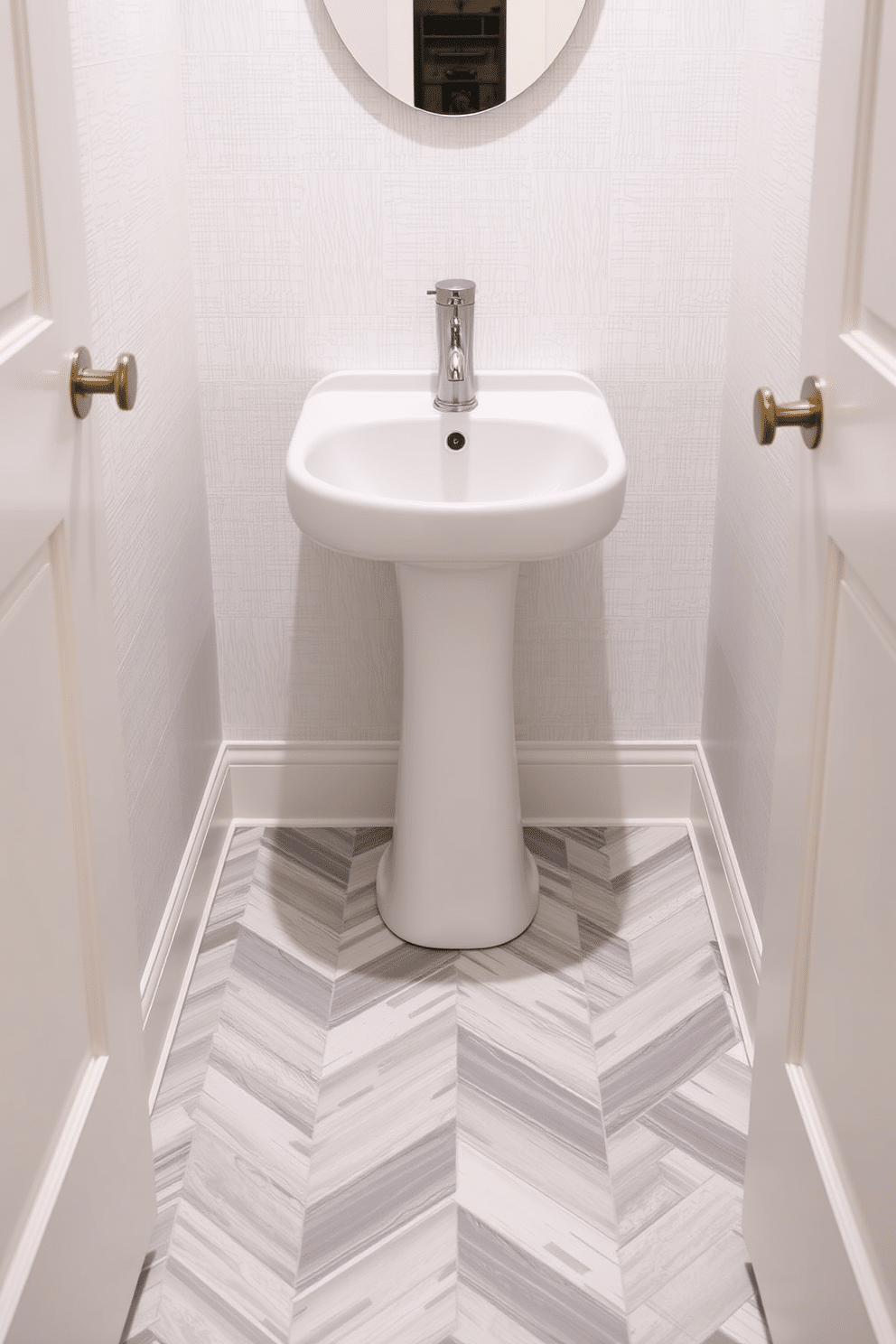 A chic powder room featuring a herringbone pattern floor tile that creates a striking visual interest. The tiles are in a mix of soft gray and white, adding a touch of elegance to the compact space. The walls are adorned with a subtle textured wallpaper in a pale hue, complementing the floor design. A sleek, modern pedestal sink is centered against the wall, accentuated by a stylish wall-mounted faucet in brushed nickel.