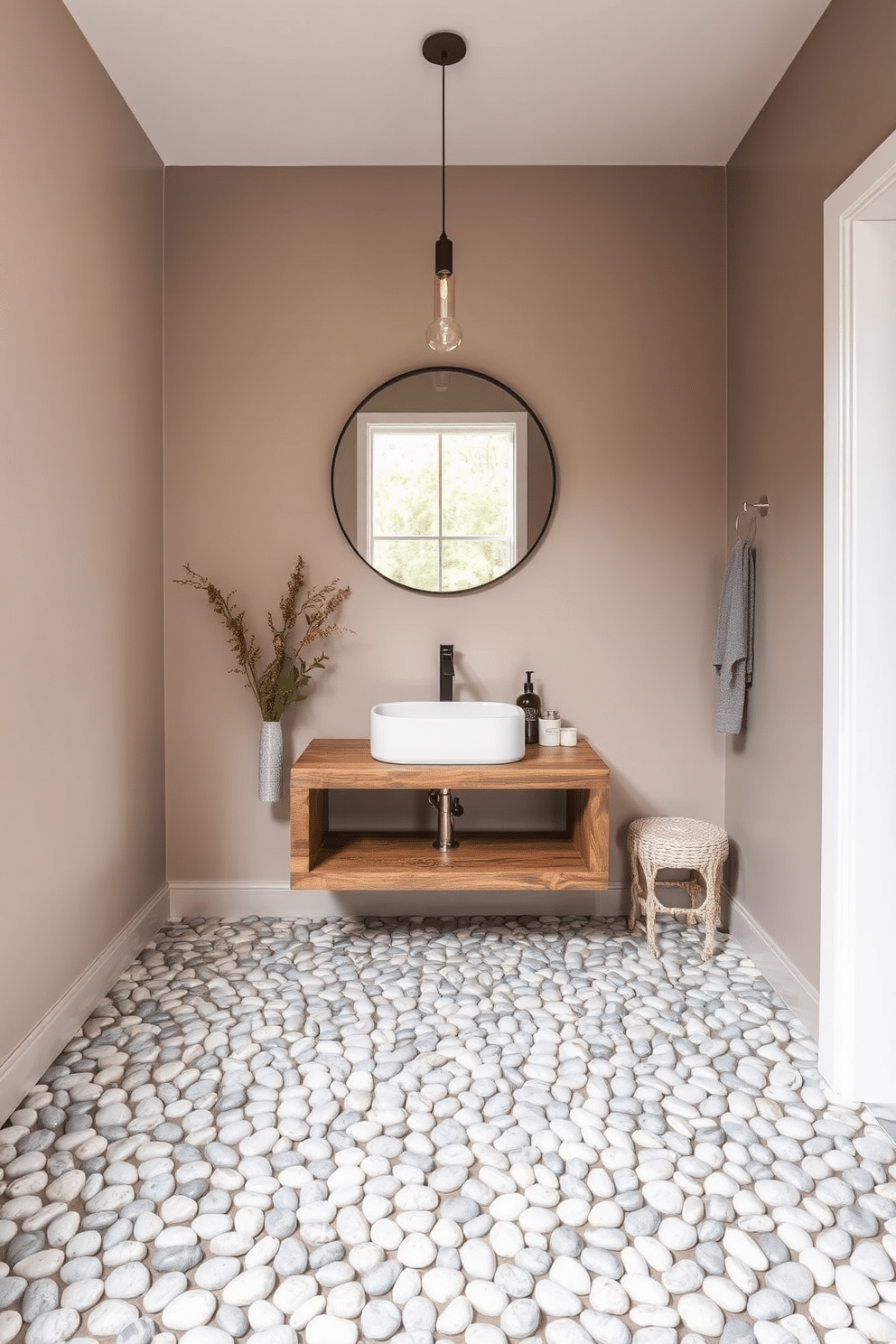 A serene powder room featuring pebble tiles that create a natural ambiance. The floor is adorned with smooth, rounded pebbles in varying shades of gray and beige, enhancing the rustic charm of the space. The walls are painted in a soft, earthy tone that complements the floor, while a sleek, modern sink sits atop a reclaimed wood vanity. A large round mirror with a minimalist frame reflects the warm light from a stylish pendant fixture overhead.