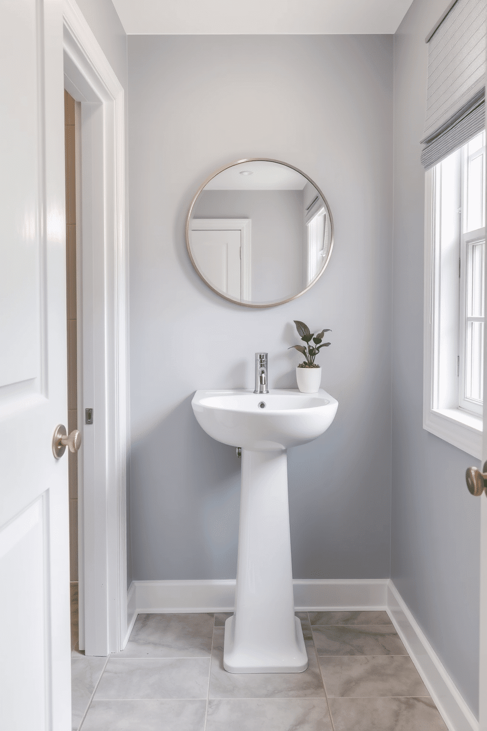 A serene powder room featuring soft gray-blue walls that create a calming atmosphere. The space includes a sleek white pedestal sink and a modern round mirror framed in brushed nickel, enhancing the tranquil vibe. The floor is adorned with light gray tiles that complement the wall color, adding to the overall elegance. A small potted plant sits on the windowsill, bringing a touch of nature into the peaceful setting.