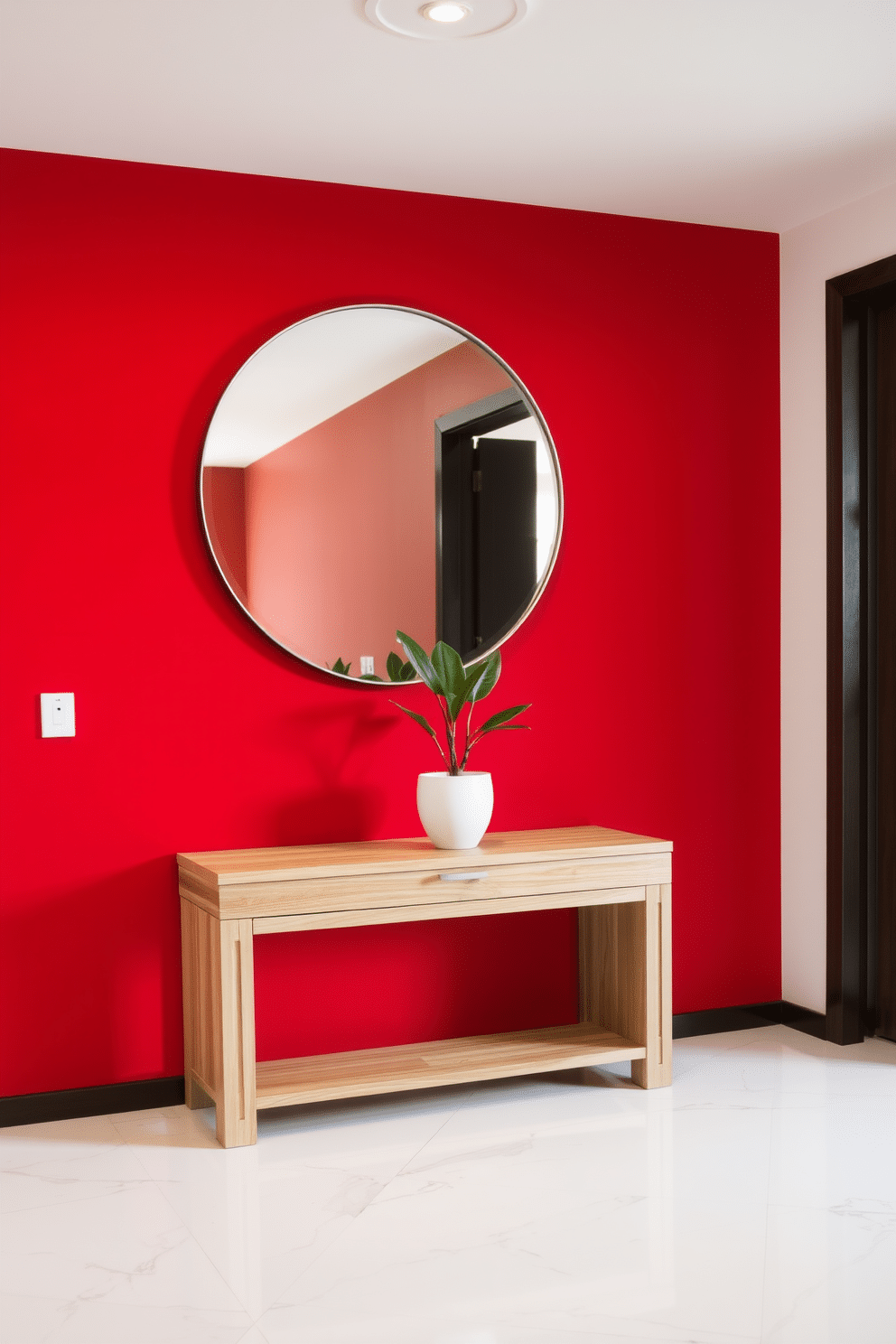 A modern minimalist foyer features a striking red accent wall that creates a bold statement. The flooring is a sleek white marble, complemented by a simple, low-profile console table in natural wood. A large, round mirror hangs above the console, reflecting the vibrant color and enhancing the sense of space. A single potted plant with clean lines sits on the table, adding a touch of greenery without overwhelming the design.