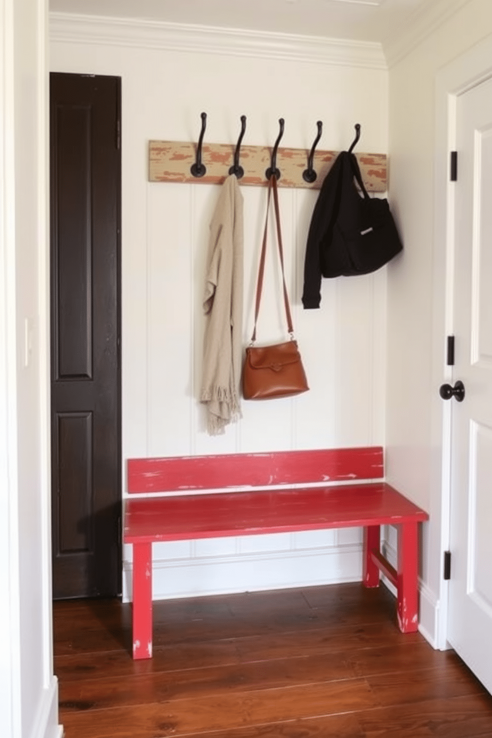 A charming farmhouse-style mudroom features a rustic red bench with a weathered finish, adorned with several hooks above for hanging coats and bags. The walls are painted in a soft cream color, complemented by a durable wooden floor that adds warmth to the space.