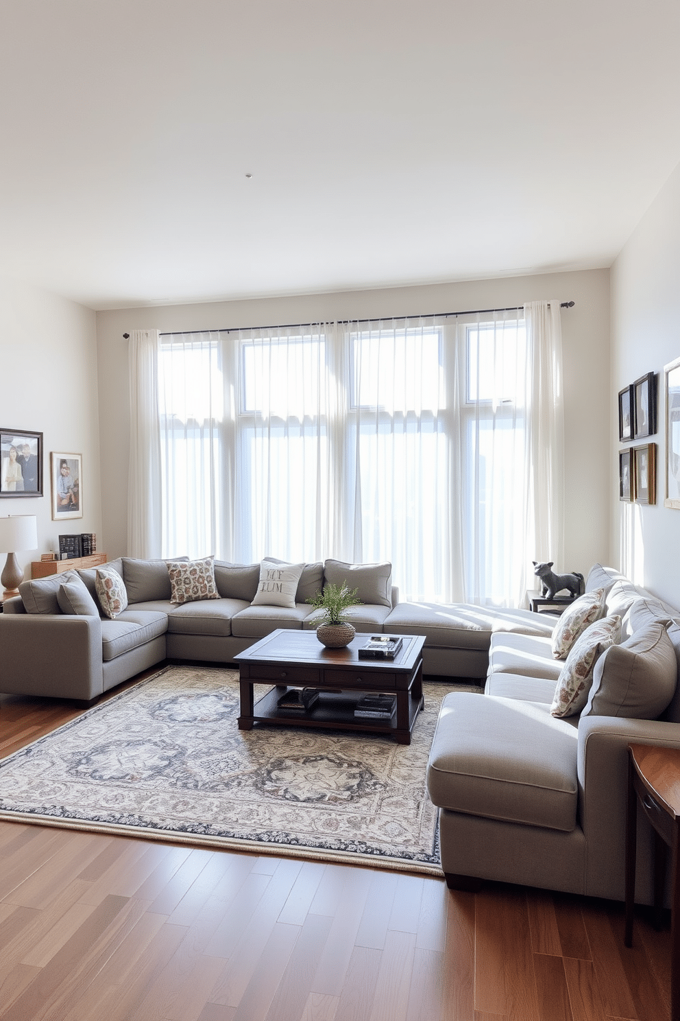 A cozy family room features a spacious sectional sofa in a soft gray fabric, arranged to create an inviting conversation area. A large area rug anchors the space, while a wooden coffee table adds warmth and functionality at the center. The walls are painted in a light beige hue, complemented by framed family photos and artwork that add personal touches. Large windows allow natural light to flood the room, with sheer curtains providing privacy while maintaining an airy feel.