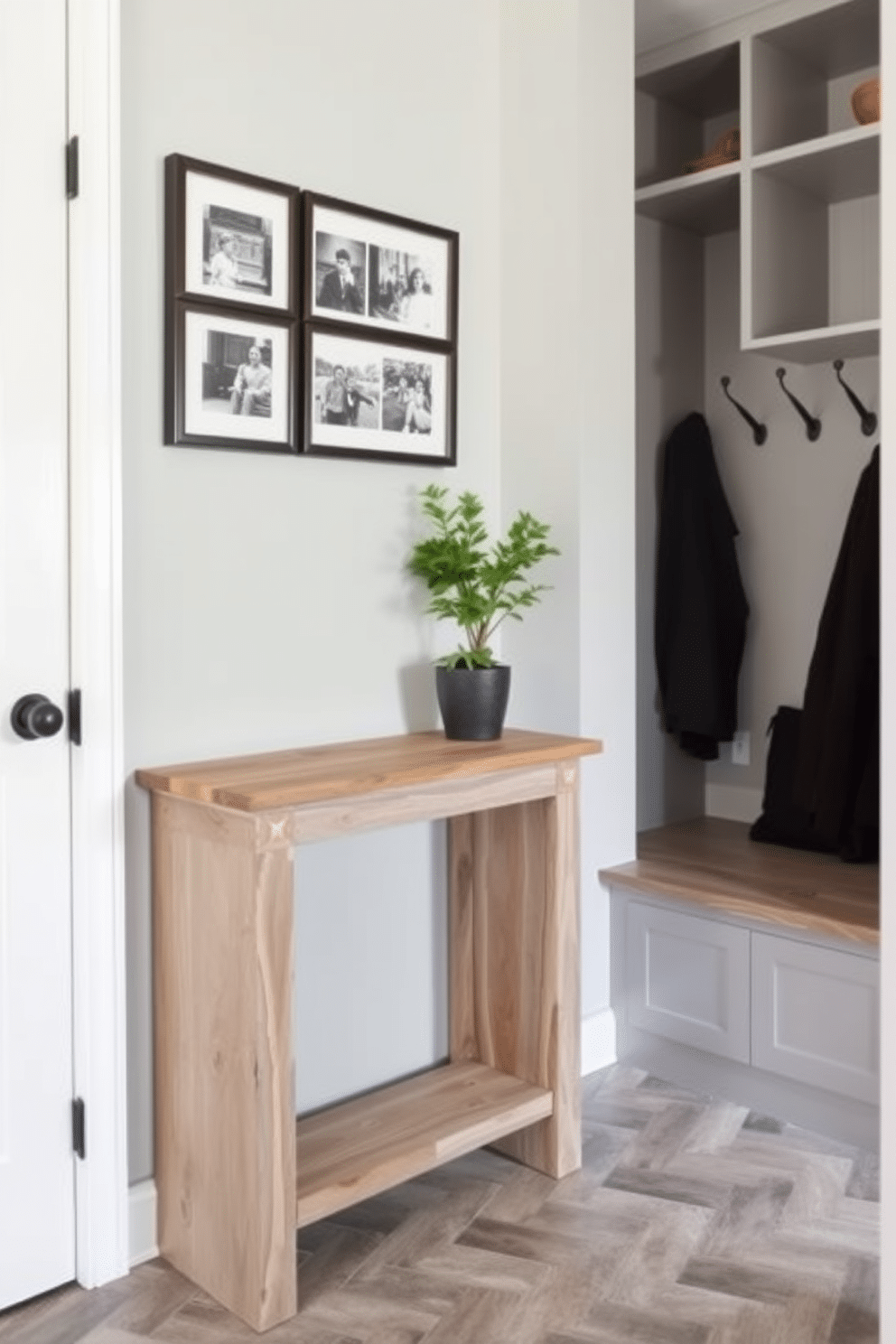 A narrow console table is positioned against the wall, crafted from reclaimed wood with a rustic finish. Above it, a series of framed black-and-white photographs adds a personal touch, while a small potted plant brings a hint of greenery. The mudroom features a practical layout with built-in benches and hooks for coats, all finished in a soft gray palette. Durable tile flooring in a herringbone pattern complements the space, making it both stylish and functional for everyday use.