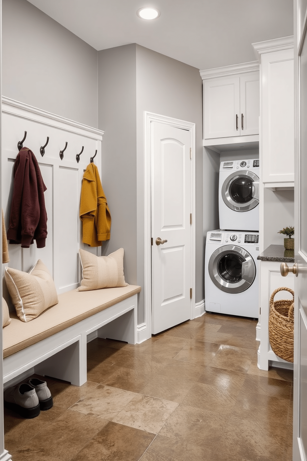 A functional mudroom connected to a laundry room features built-in storage benches with plush cushions and hooks for coats. The walls are painted in a soft gray, and the floor is adorned with large, durable tiles that can withstand heavy foot traffic. Adjacent to the laundry area, a compact washer and dryer are seamlessly integrated into cabinetry, providing a clean and organized look. A small potted plant and a woven basket for shoes add a touch of warmth and style to the space.