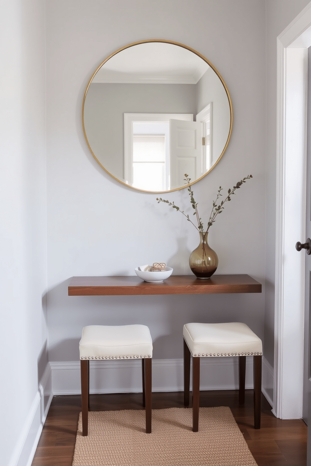 A narrow console table made of sleek, dark wood stands against a light gray wall, complemented by two elegant, upholstered stools in a soft cream fabric. Above the table, a large round mirror with a thin gold frame reflects natural light, enhancing the space's brightness and inviting ambiance. On the console table, a small decorative bowl holds keys and a few stylish coasters, while a slender vase with fresh greenery adds a touch of life. The floor is adorned with a textured runner that ties the color scheme together, creating a warm and welcoming entryway.