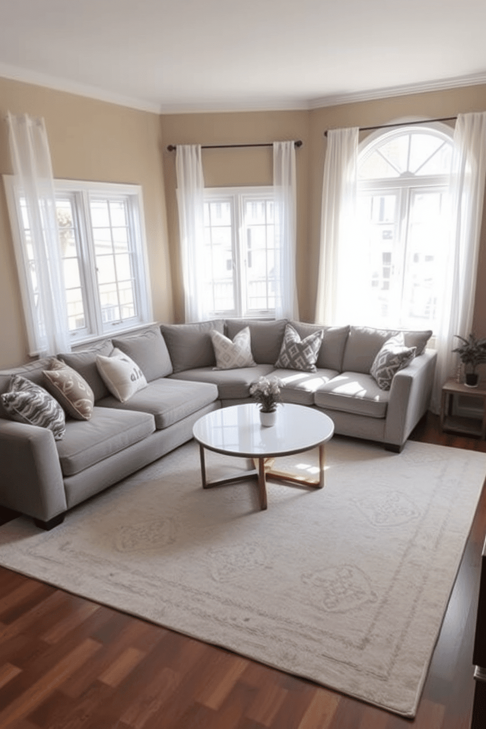 A cozy small family room featuring a round coffee table at the center, surrounded by a plush sectional sofa in a soft gray fabric. The walls are painted in a warm beige, and a large area rug with subtle patterns anchors the space, adding texture and comfort. Natural light floods the room through large windows adorned with sheer white curtains, creating an inviting atmosphere. Decorative throw pillows in various patterns and colors are scattered across the sofa, enhancing the room's playful yet sophisticated vibe.