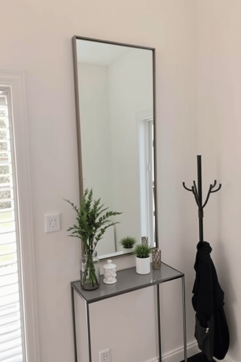 A functional mirror is mounted on the wall of a small foyer, featuring a sleek frame that complements the overall decor. Beneath the mirror, a narrow console table holds decorative items and a small potted plant, adding a touch of greenery to the space. The walls are painted in a soft, neutral tone, enhancing the natural light that floods in through the nearby window. A stylish coat rack stands to one side, providing a practical solution for hanging outerwear while maintaining an organized look.