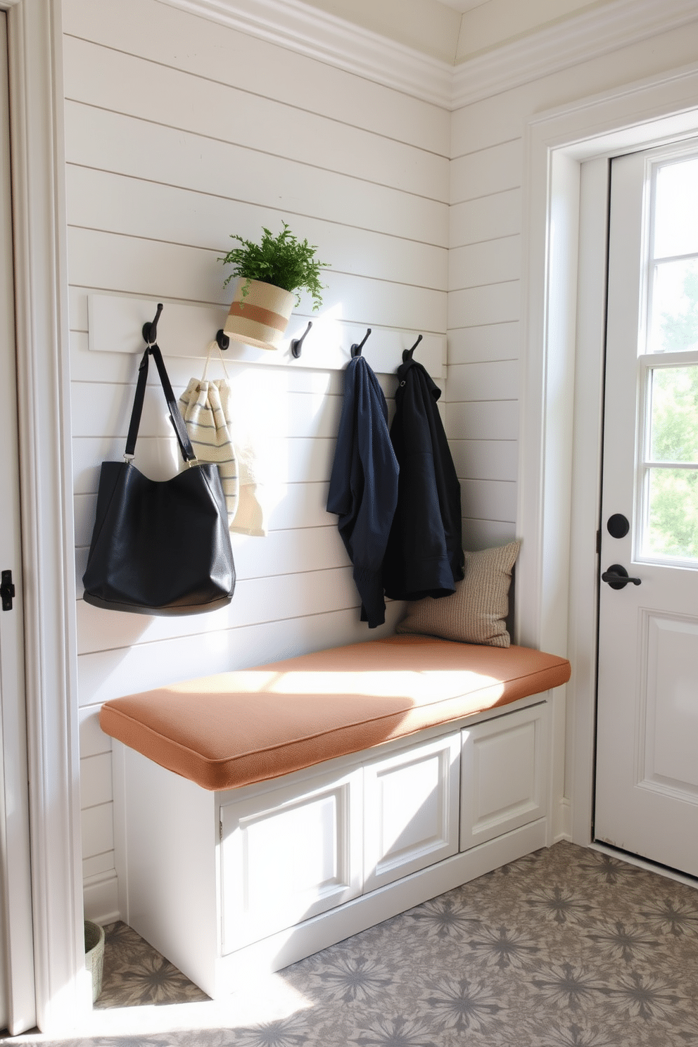 A cozy bench with hidden storage is placed against the wall, featuring a soft, cushioned top in a warm, neutral fabric. Above the bench, there are hooks for hanging coats and bags, and a small potted plant adds a touch of greenery. The mudroom is designed with a light color palette, incorporating shiplap walls and a durable, patterned tile floor. Natural light streams in through a nearby window, illuminating the space and enhancing its inviting atmosphere.