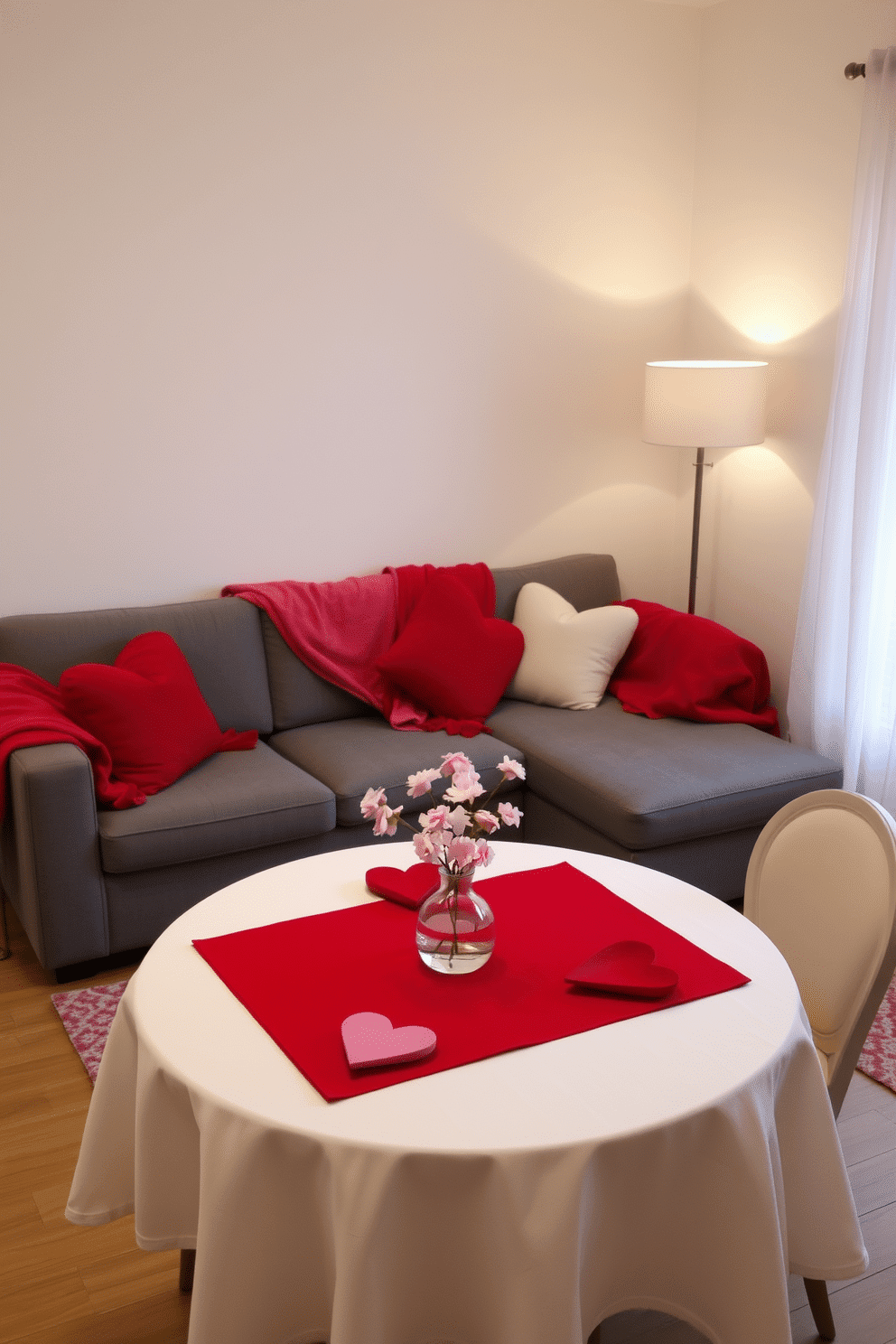 A cozy living room adorned with red and pink throw blankets draped over a plush gray sofa, creating a warm and inviting atmosphere. Soft, ambient lighting from a nearby floor lamp enhances the romantic feel, while heart-shaped cushions add a festive touch. In a small dining nook, a round table is set for two, featuring a simple white tablecloth topped with a vibrant red runner. Delicate pink flowers in a glass vase serve as a centerpiece, complemented by heart-themed place settings that celebrate Valentine's Day.