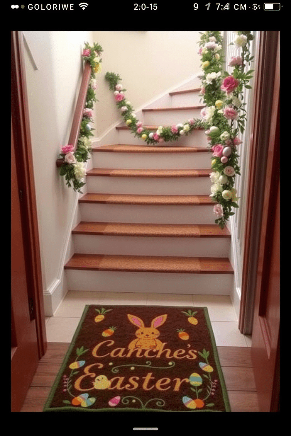 A charming Easter-themed doormat welcomes guests at the bottom of the staircase, featuring pastel colors and playful bunny motifs. The staircase is adorned with garlands of spring flowers and colorful eggs, creating a festive atmosphere that invites warmth and cheer.