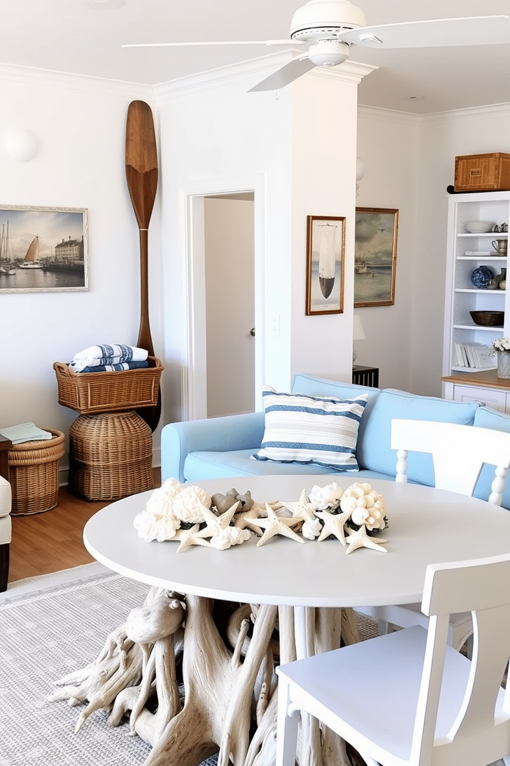 A bright and airy summer apartment filled with nautical decor. The living room features a light blue sofa adorned with striped throw pillows and a large seashell centerpiece on the coffee table. In the corner, a vintage wooden boat oar is mounted on the wall above a woven basket filled with beach towels. The dining area showcases a round table with a driftwood base, surrounded by white chairs and a centerpiece of starfish and coral.