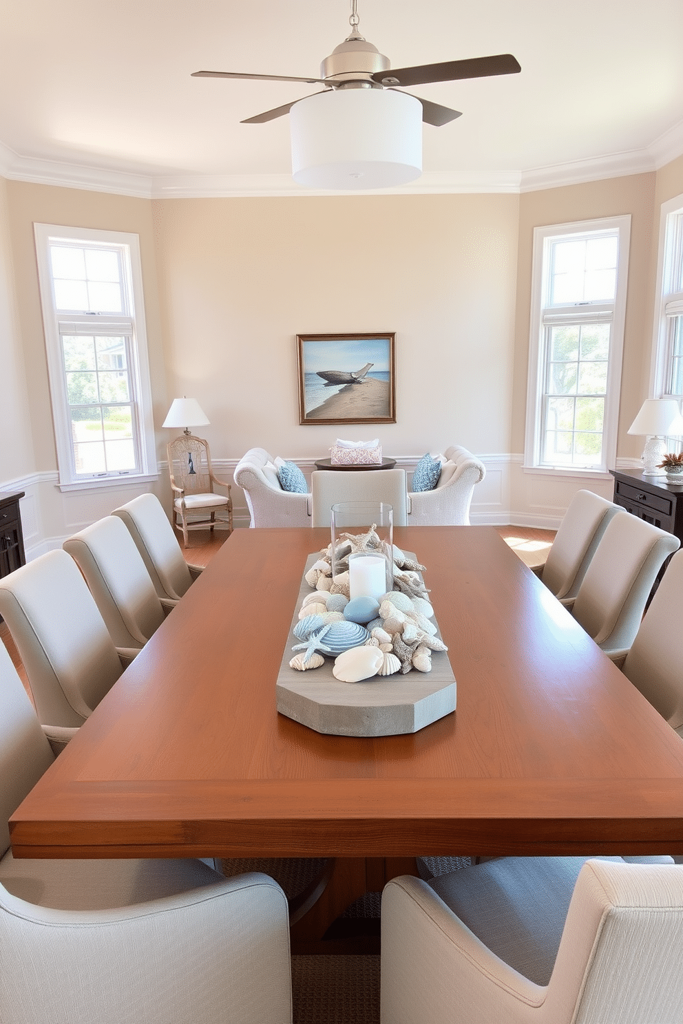 A bright and airy dining room features a large wooden table surrounded by comfortable upholstered chairs. At the center of the table, a coastal-themed centerpiece is displayed, incorporating seashells, driftwood, and soft blue accents. The walls are painted in a light sandy beige, complementing the natural textures throughout the space. Large windows allow plenty of sunlight to filter in, enhancing the cheerful summer atmosphere.